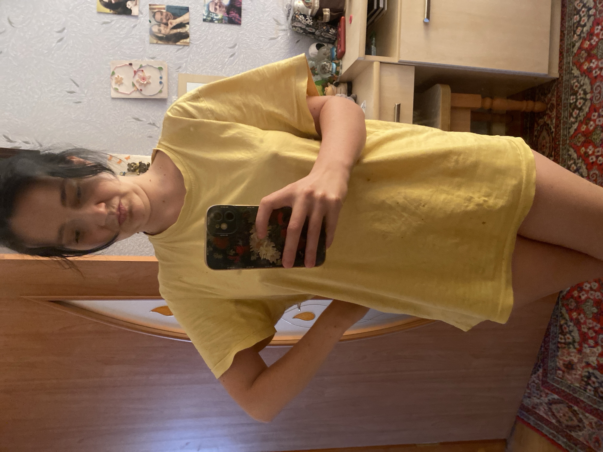 A young woman with dark hair is reflected in a mirror, holding a smartphone in her left hand. She is wearing a yellow t-shirt and appears to be in a bedroom. Behind her, a wooden door and a wall with several framed photographs are visible. A dresser and a small stool are also in the background. The floor is covered with a patterned rug.
