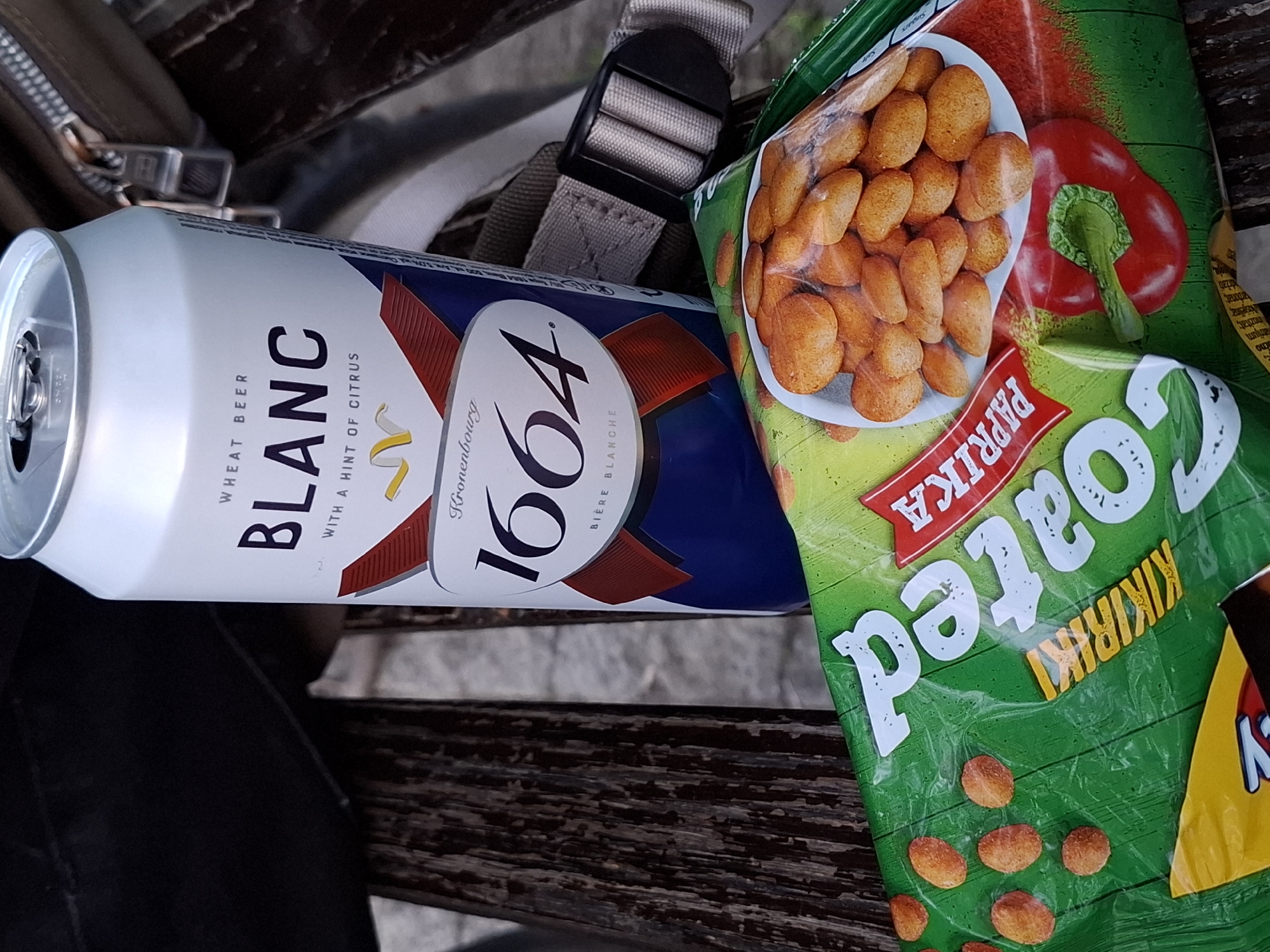 An eye-level shot captures a can of beer and a bag of snacks resting on a weathered wooden surface, likely a bench or table. The setting appears to be outdoors in Novi Sad, Serbia.
The central object is an open white can of "1664 BLANC" wheat beer, which also states "WITH A HINT OF CITRUS", "Kronenbourg", and "BIÈRE BLANCHE". Part of its "OLD" text is also visible. Adjacent to the can is a green bag of "Coated PAPRIKA KIKIRIKI" (peanuts), prominently displaying a red bell pepper illustration and several of the orange-coated peanuts. The bag also has text in Serbian, including "trijum", "arbonat", and "držati" (meaning "keep"), along with "inozitat" and "Sugars" in English.
In the background, a beige strap with a black buckle and a brown bag or backpack with a zipper are partially visible, suggesting these items belong to someone taking a break. The wooden surface beneath the items has a rich texture with visible grain and wear. The natural lighting indicates it is daytime.