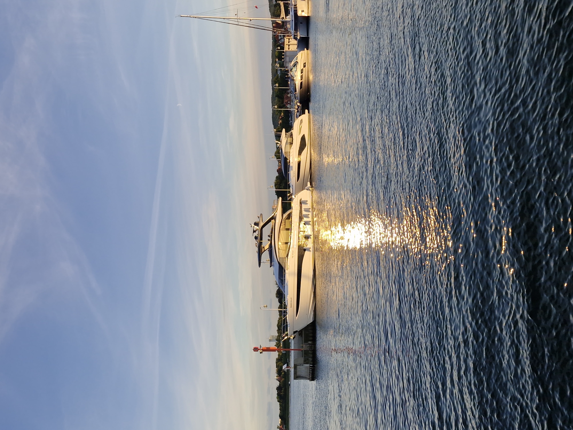 The image depicts a harbor in Sopot, Poland, at sunset. Several modern yachts are docked along a pier. The water is calm, with a golden reflection of the setting sun shimmering on its surface. In the background, a line of trees and buildings forms the coastline. The sky is a clear blue with a few wispy clouds, and a crescent moon is faintly visible. A red and white navigational buoy is situated at the end of the pier, to the left of the yachts.