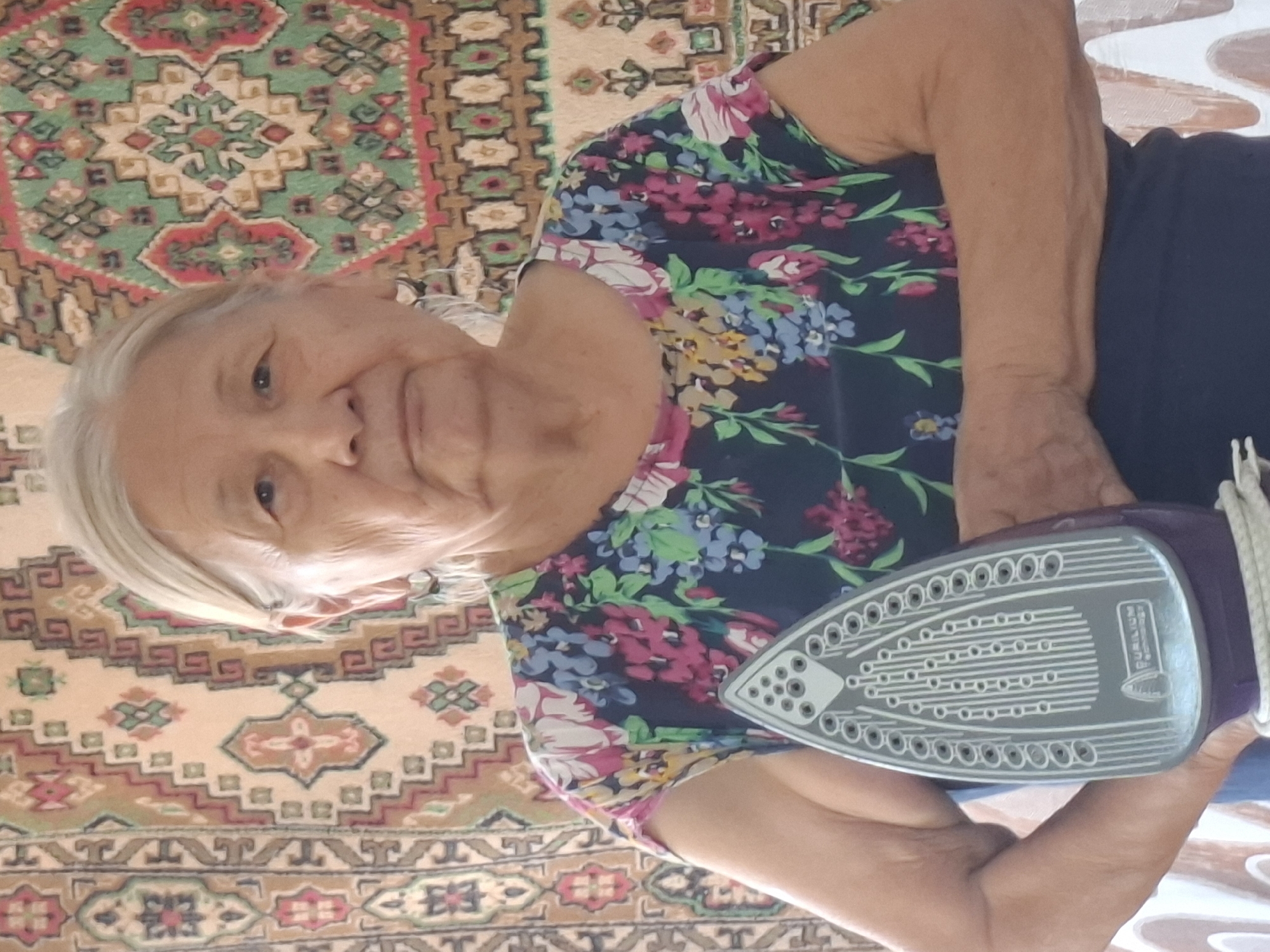An elderly woman with grey hair and fair skin is shown from the chest up. She is wearing a dark blue t-shirt with a floral pattern of pink, blue, and yellow flowers. She is holding a purple iron in her right hand and her left arm is bent, resting on her chest. The background is a tapestry with a geometric pattern of red, green, and beige. The image appears to have been taken indoors, possibly in Almaty, Kazakhstan.