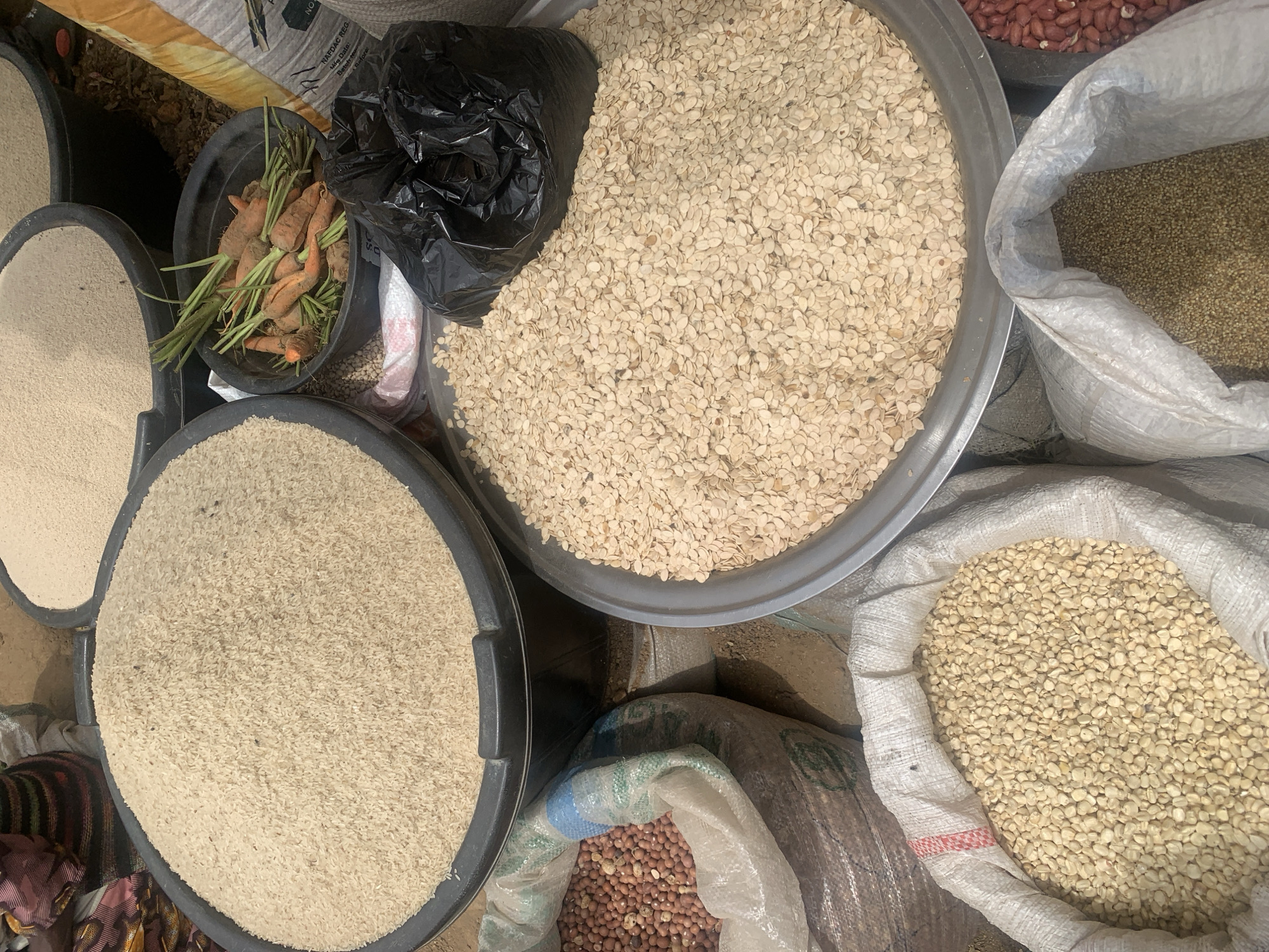 The image depicts a marketplace stall in Jos, Nigeria. Various food items are displayed in containers. There are round, flat trays containing rice, and a light-colored grain. A black bucket contains a handful of small carrots with green tops, partially obscured by a black plastic bag. Bags hold peanuts, a small grain, and white beans. The setting suggests a local market environment.