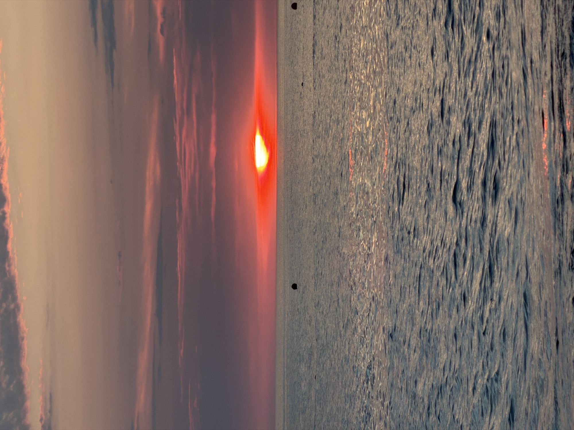 The image depicts a sunset over the Black Sea, likely near Sochi, Russia, given the typical weather patterns and coastal features associated with the region. The sun, a brilliant orange-yellow orb, is partially obscured by a band of clouds near the horizon, casting a warm, reddish glow across the sky. Wispy clouds, tinged with pink and purple, stretch across the upper portion of the frame. The sea is calm, with gentle ripples reflecting the colors of the sky. In the distance, a few dark buoys are visible on the water's surface. The overall atmosphere is serene and picturesque, typical of a coastal sunset.