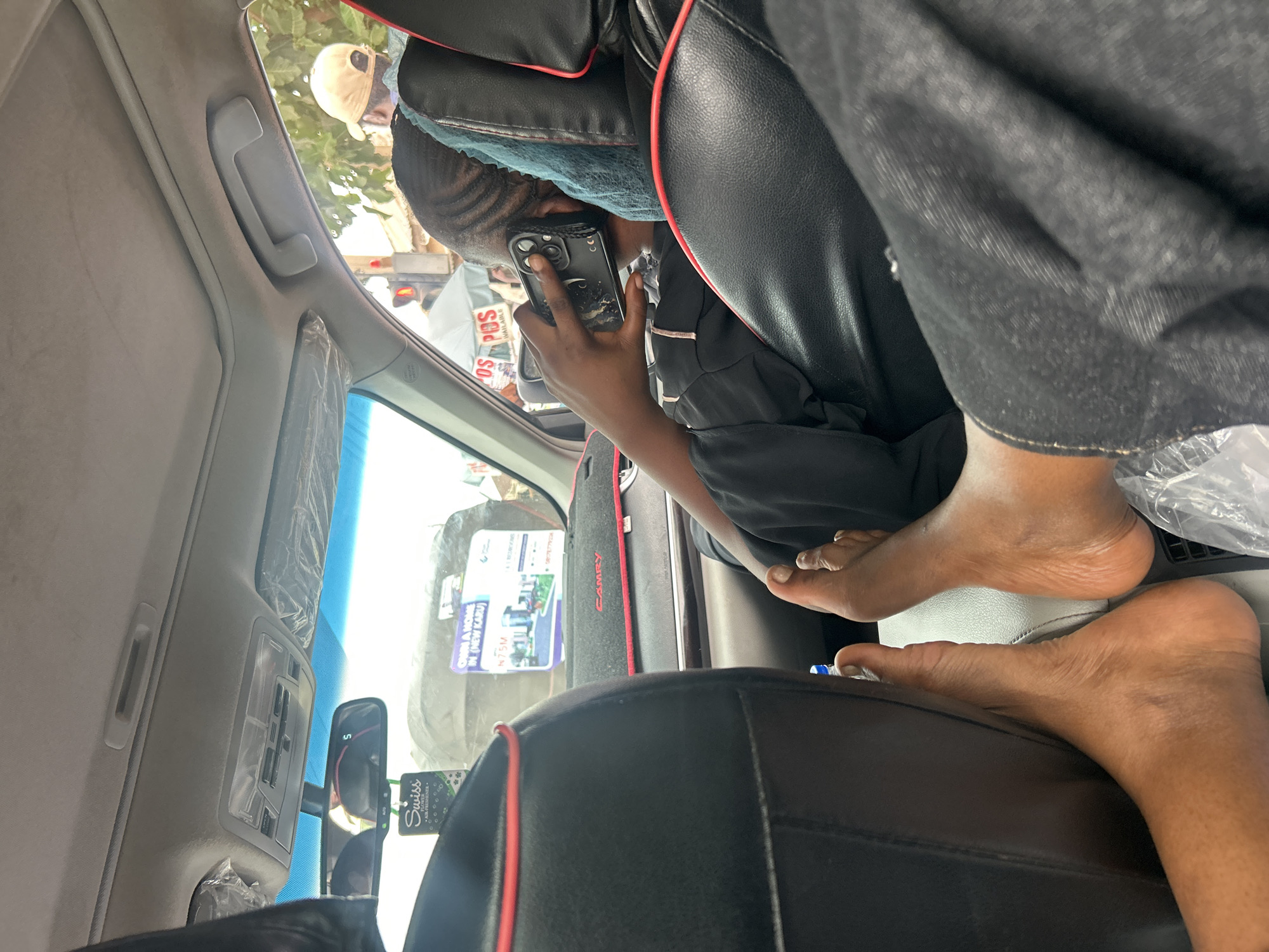 The media file shows the interior of a car, likely in Karu, Nigeria, with several people and objects visible. The camera angle is tilted, and the scene appears to be captured during the daytime, with bright lighting suggesting clear weather.
In the foreground, two pairs of bare feet are visible, resting on what appears to be car seats. One pair of feet belongs to a person sitting in the back seat, with their legs and feet exposed. The other pair is positioned further back, possibly from another passenger.
In the middle ground, a person is seated in the back of the car, holding a smartphone to their ear as if on a call. They are wearing dark clothing and have a light-colored cloth draped over their head. A glimpse of a person wearing a beige baseball cap is visible through the car window, suggesting they are outside the vehicle.
The car interior features black leather seats with red trim and a gray headliner. Various items are attached to the car's interior, including a "Swiss Air Freshener" hanging from the rearview mirror. Visible text on a placard inside the car reads "OWN A HOME IN (NEW KARU) N75M" and "POS AVAILABLE," indicating real estate and financial service advertisements. The word "CAMRY" is visible on a trim piece of the car.
The overall scene depicts a typical moment inside a vehicle, possibly during a commute or a journey, with passengers engaged in personal activities and the presence of advertisements common in many urban environments. The context of Karu, Nigeria is provided by the visible text on the advertisement.
