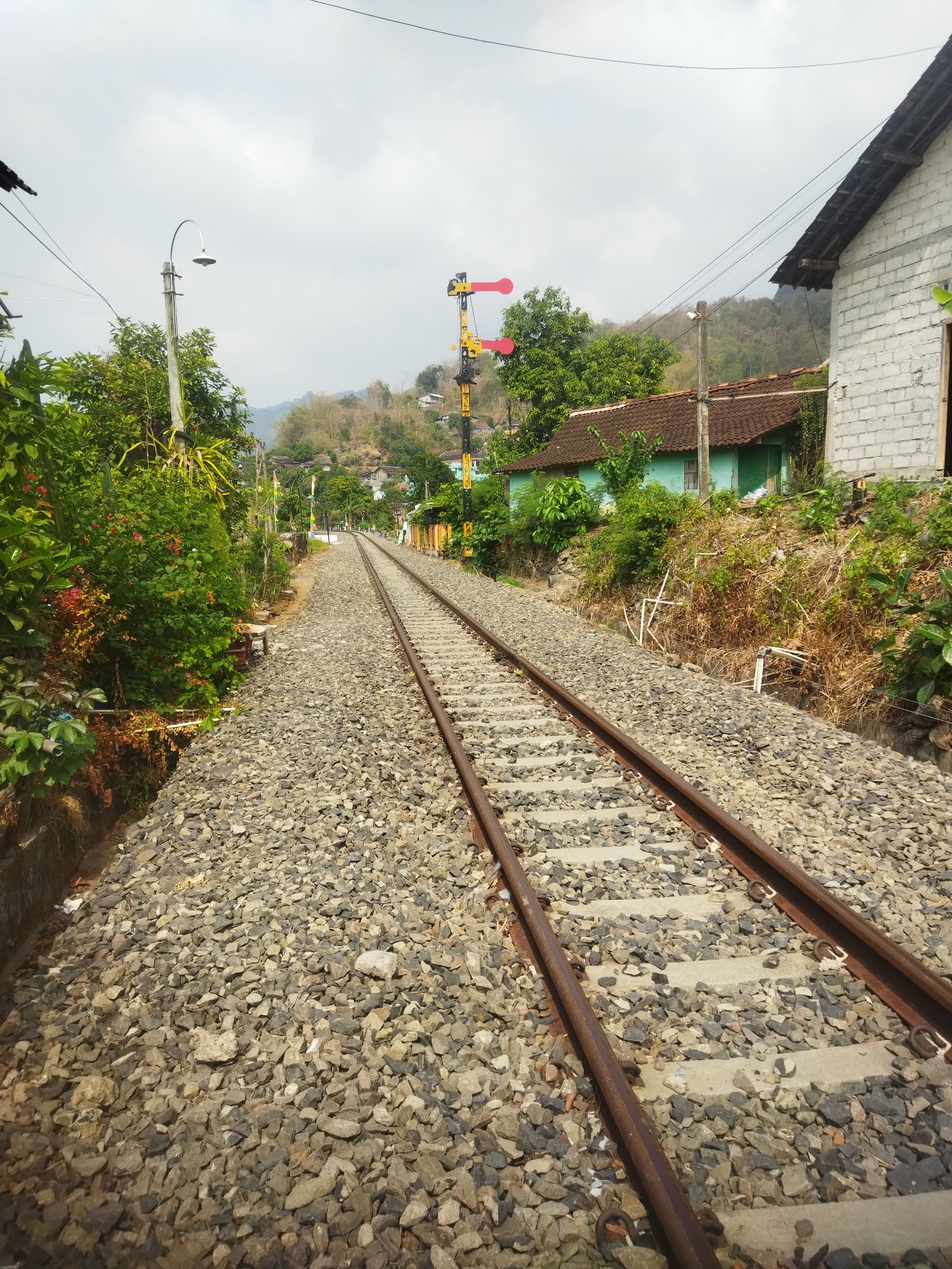 The image captures a view of a single-track railway line in the city of Wonogiri, Indonesia. The tracks are laid on ballast composed of gray and brown stones, with concrete sleepers visible beneath the rails. The railway extends into the distance, curving slightly to the left.
On the left side of the tracks, dense green foliage and bushes with some red flowers are present. A concrete wall or embankment runs alongside the tracks. A lamppost with a curved arm and a light fixture stands tall amongst the greenery.
On the right side of the tracks, a white brick building with a brown tiled roof is prominently featured. Adjacent to it is a smaller, light blue house with a dark brown tiled roof and green doors and windows. The area surrounding the houses is covered in dry grass and vegetation.
Further down the track, a railway signal with three colored lights (red, yellow, and red) is visible on a yellow pole, indicating the status of the railway line. In the background, rolling hills covered with trees and some scattered houses suggest a semi-rural or suburban setting. The sky is overcast with a pale, cloudy appearance. The scene depicts a tranquil, everyday view of a functional railway in a populated area of Wonogiri.