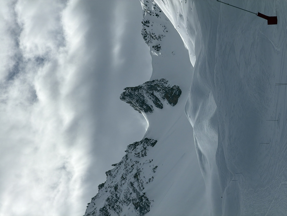 The image captures a snow-covered mountainous landscape in Courchevel, France, under an overcast sky. A prominent, rugged rock pinnacle, partially covered in snow, dominates the central mid-ground, flanked by extensive snow-clad slopes and jagged peaks. The foreground consists of undulating snowfields featuring numerous ski tracks. Several thin, dark poles are sparsely distributed across the lower-left slope, potentially marking a trail. On the far right, a red, angular object, possibly the base of a snow cannon or a ski lift support structure, is partially visible. The sky is predominantly grey with patches of white clouds, indicating diffused light conditions. No individuals or specific actions are discernible.