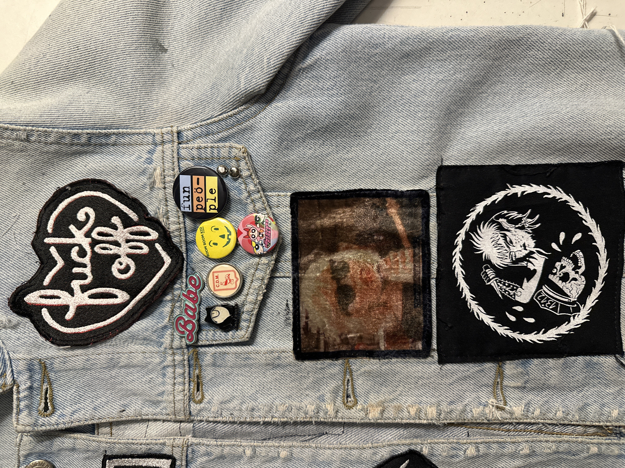 The image shows a section of a light-wash denim jacket. Patches and pins are attached to the jacket. From left to right: a patch with a white design on a black background with red trim, pins including a "fun people" pin, a yellow smiley face pin with "boom boom kid" written on it, a pin with a cartoon character, a "C.D.M." pin, a pin with a penguin, and a "Babe" pin. Next is a square patch with a mottled brown and black design, and then a black square patch with a white circular design featuring a dragon and skull. The background is a neutral surface.