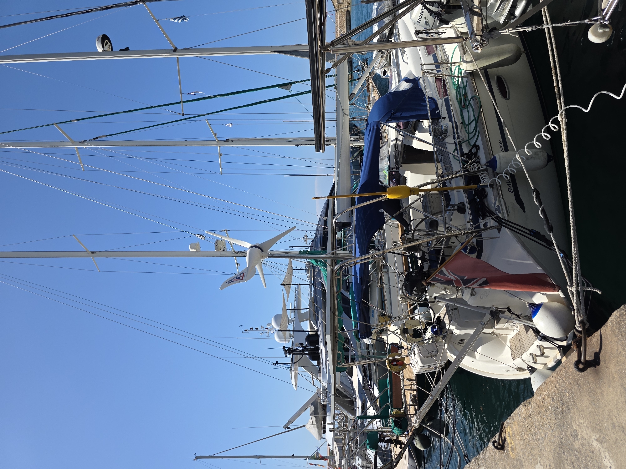 In the harbor of Chania, Greece, on a bright, clear day with a cerulean sky, a fleet of sailboats and yachts are moored. The foreground features a white sailboat with a blue canopy, a red British flag, and a white coiled rope tethering it to a concrete dock. White buoys are visible along the hull. Behind it, other sailboats and larger yachts, including a white luxury yacht with multiple decks and a black radar dome, are docked. A white wind turbine with three blades is mounted on the mast of one of the sailboats. The water is dark green and calm.