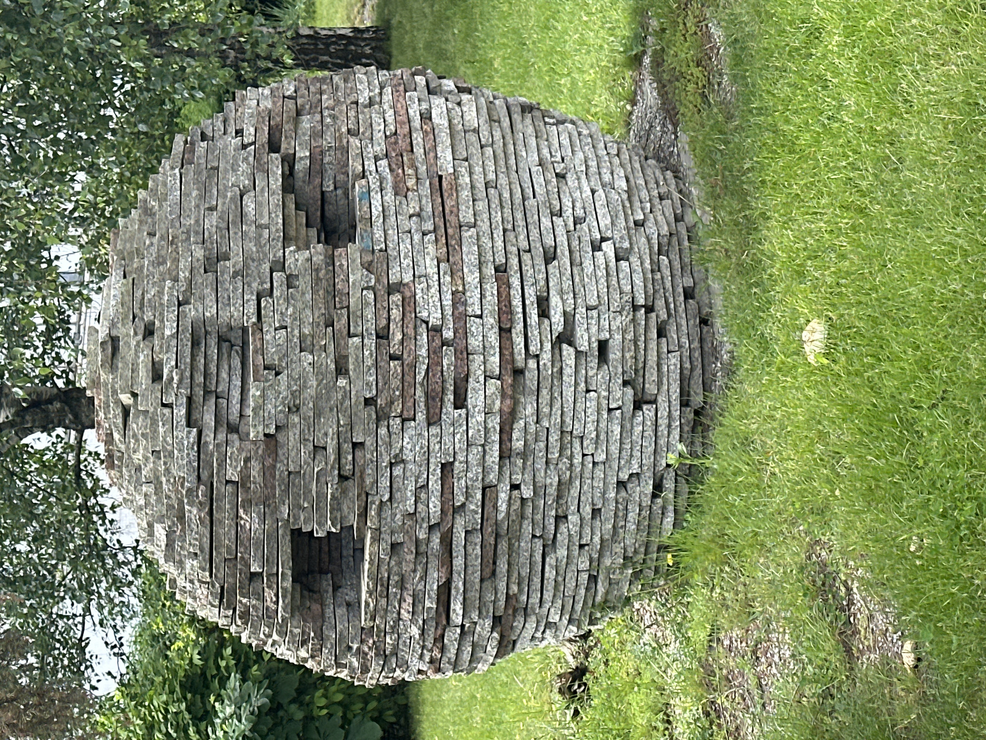 The media file captures a large, spherical sculpture positioned outdoors in what appears to be a park or garden in Minsk, Belarus. The sculpture is constructed from numerous rectangular, flat stone slabs, stacked horizontally and varying in length, creating a textured and segmented surface. The stones are predominantly grey, with some interspersed reddish-brown pieces contributing to a varied color palette. Two circular indentations or openings are visible on the side of the sphere facing the viewer.
The sculpture rests directly on a vibrant green lawn, which fills the lower and right portions of the frame. In the background and to the left, leafy green trees are visible, suggesting a natural, vegetated environment. The lighting indicates it is daytime, with bright, even illumination suggesting clear or partly cloudy weather. There are no people visible, and no specific activity or event is taking place, presenting a static scene focused on the artwork and its natural surroundings. No visible text is present in the image.