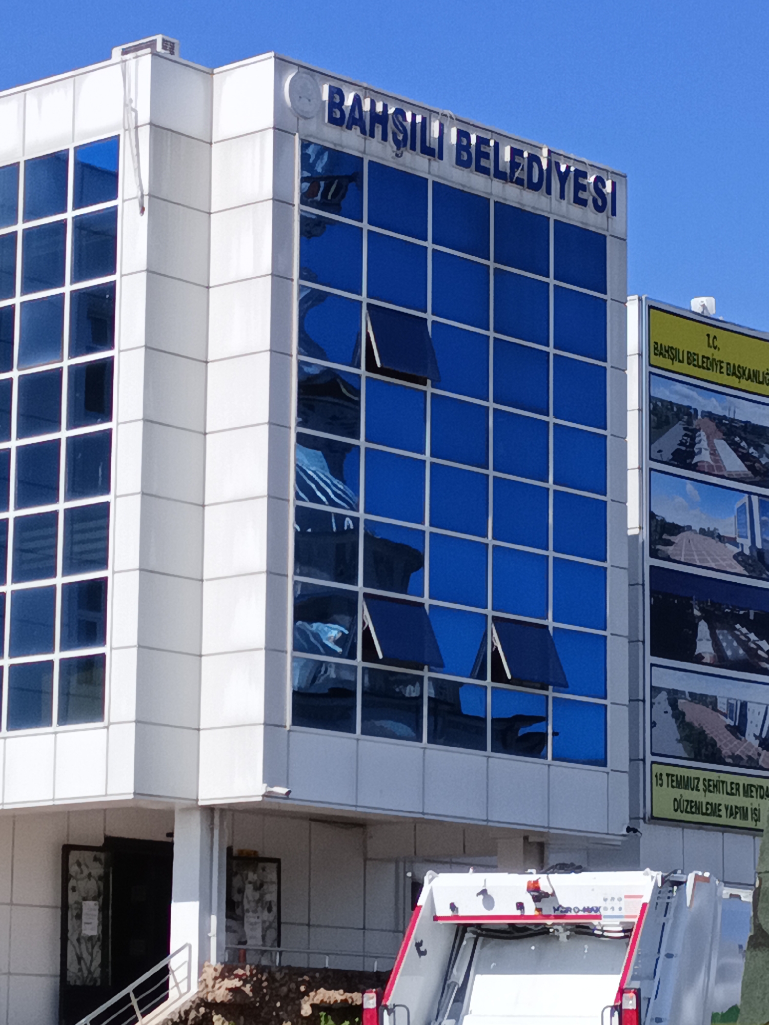 The image shows the exterior of the Bahşılı Belediyesi (Bahşılı Municipality) building in Turkey. The building features a modern white facade with large blue-tinted glass windows. The words "BAHŞILI BELEDİYESİ" are visible in large blue letters on the top of the building. To the right, a yellow sign with black text reads "T.C. BAHŞILI BELEDİYE BAŞKANLIĞI" (Republic of Turkey Bahşılı Municipality Directorate). Below this, there are several framed images or posters showcasing urban development projects, including one that reads "15 TEMMUZ ŞEHİTLER MEYDANI DÜZENLEME YAPIM İŞİ" (15 July Martyrs Square Arrangement Construction Work). In the foreground, part of a white garbage truck with red accents is visible. The sky is clear and blue.