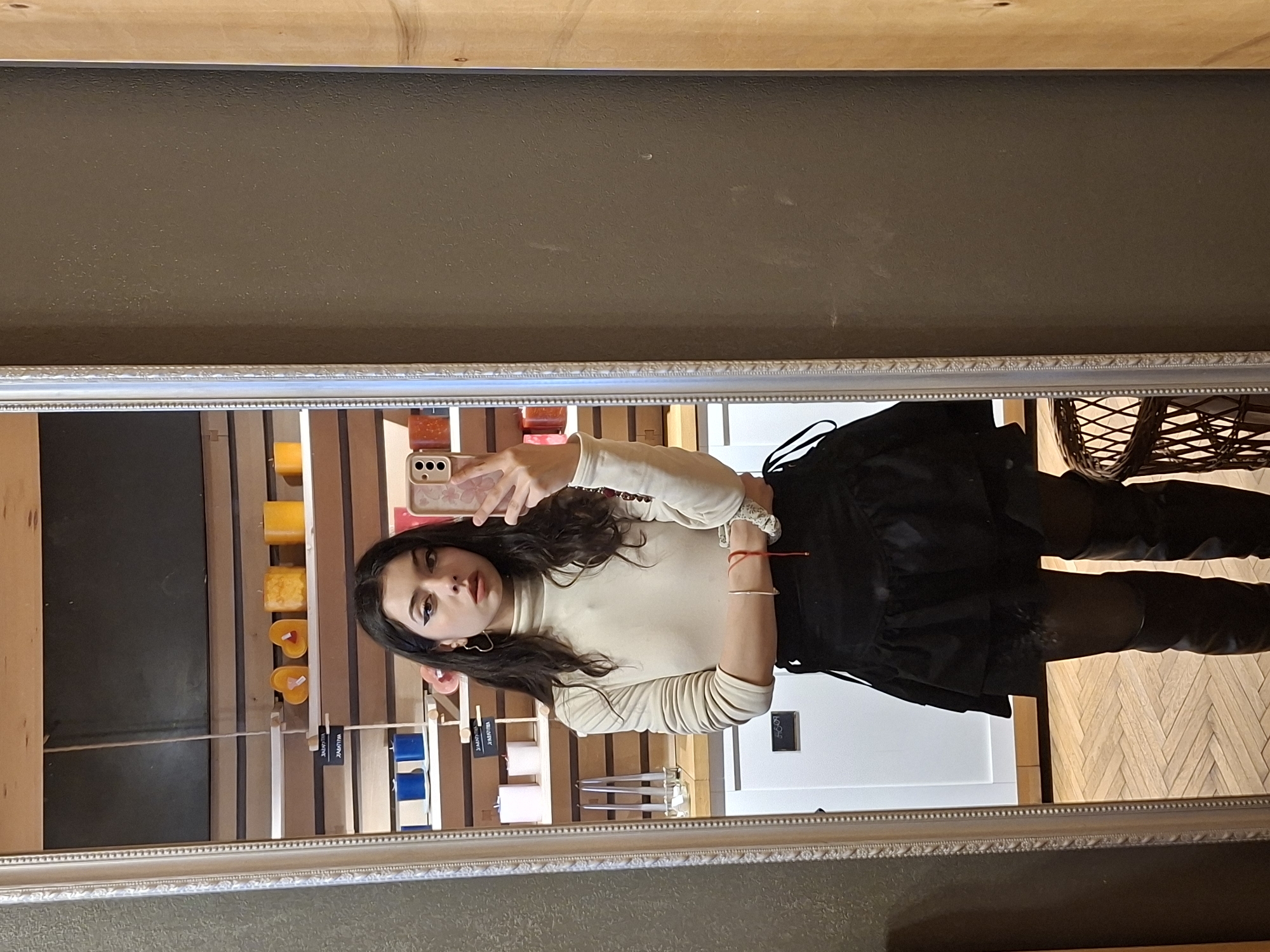 A mirror selfie shows a person with dark hair, wearing a light-colored turtleneck, a black skirt, and black leggings. They are holding a phone in their hand, taking the picture in what appears to be a store. Candles and other merchandise are visible in the background. The location is in the city of Unknown, Unknown.