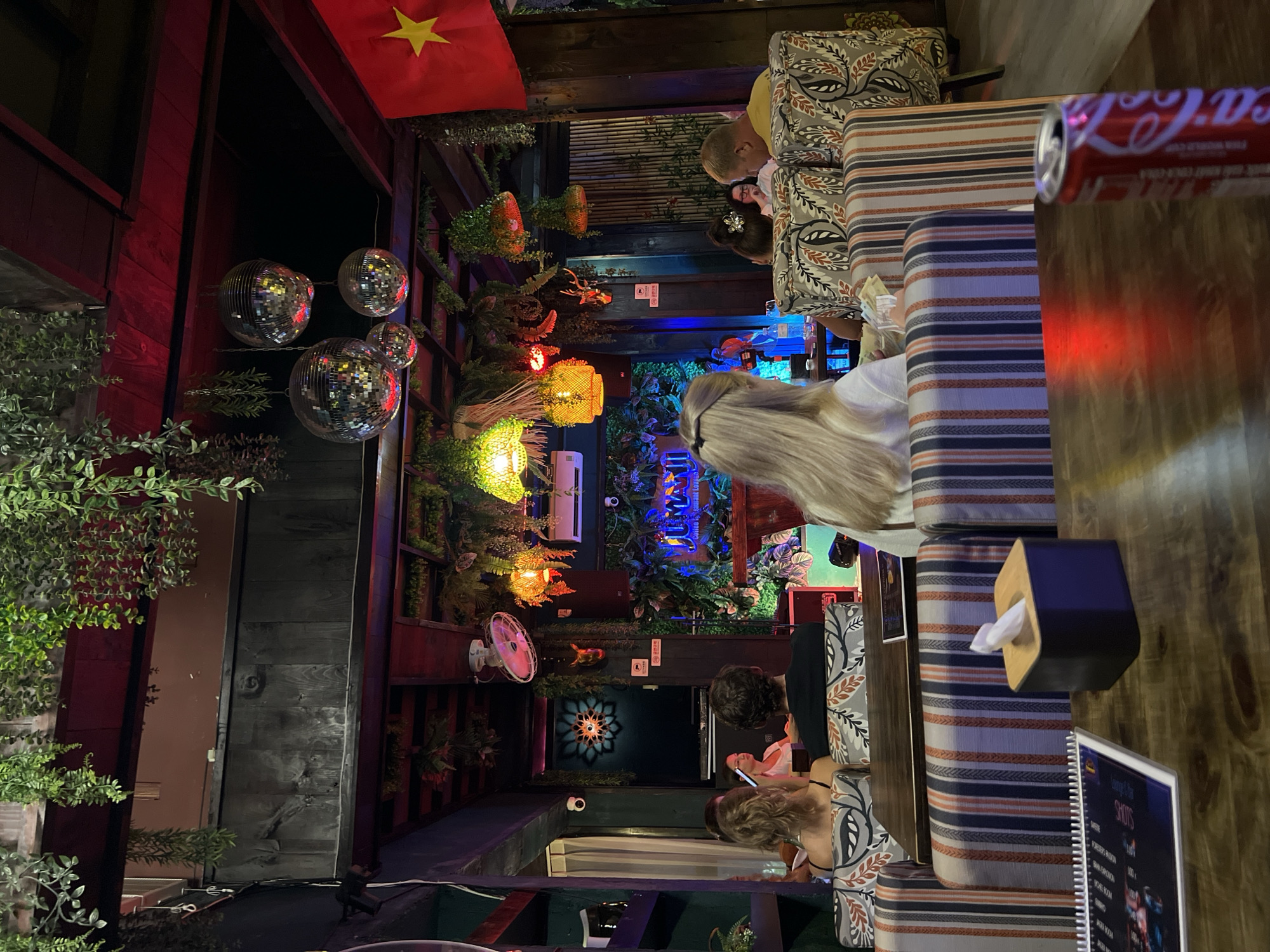 Here's a detailed description of the image:
**Setting:** The image appears to be taken inside a bar or restaurant at night. The location is unidentified, indicated by "Unknown, Unknown". The interior design is eclectic, with a mix of modern and traditional elements.
**People:** There are several people visible in the scene. Some are seated at tables, possibly dining or having drinks. There's interaction between some of the individuals, suggesting they are in conversation or socializing. Specific details such as age, gender, and clothing are hard to discern due to the angle and lighting.
**Objects:**
* **Lighting:** Decorative lighting is a prominent feature, including string lights, lanterns, and disco balls, creating a vibrant atmosphere.
* **Decor:** There are several decorative elements like plants, both real and artificial, and a Vietnamese flag.
* **Furniture:** Striped and patterned upholstered seating is visible.
* **Table items:** A Coca-Cola can, a tissue box, and a menu can be seen on the tables.
* **Signage:** There is signage, possibly advertising or indicating the name of the establishment.
* **Fan:** There is a white fan mounted on the wall.
**Actions & Interactions:**
* People are sitting and socializing.
* Some individuals seem to be using their phones.
**Environment:** The environment is dark and somewhat cramped. There's a mix of textures and materials, including wood, metal, and fabric. The overall atmosphere is lively and festive.