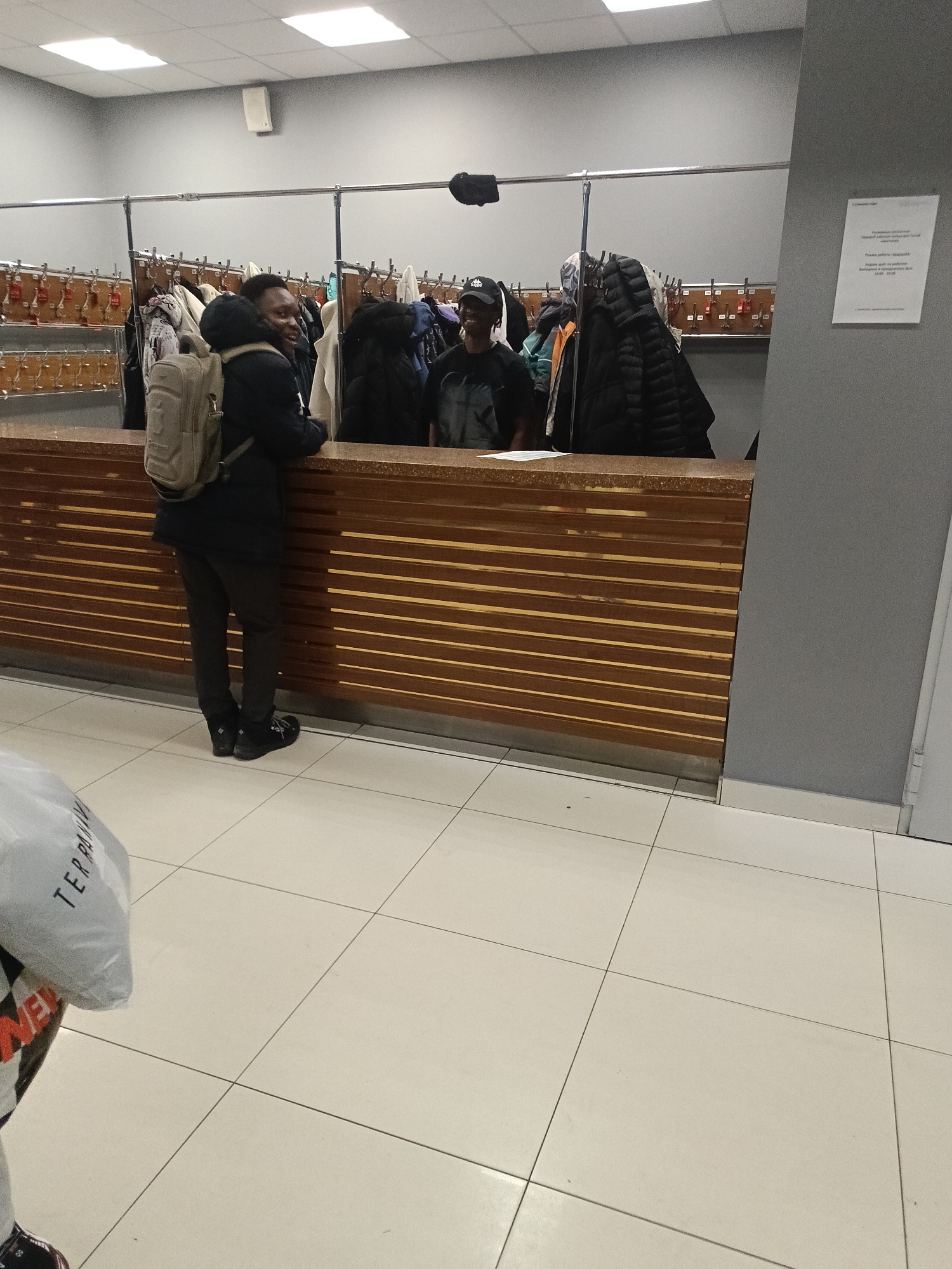 The image shows an indoor coat check area. Two men are visible behind a wooden counter. The man on the left wears a dark jacket and pants with a tan backpack. The man on the right wears a dark t-shirt and a black cap. Behind the counter, there's a row of hanging coats and a rack with numbered keys. A sign is posted on the wall to the right. The floor is tiled, and a shopping bag is visible in the lower left corner. It is the coat check at the Talisman shopping center in Izhevsk, Russia.