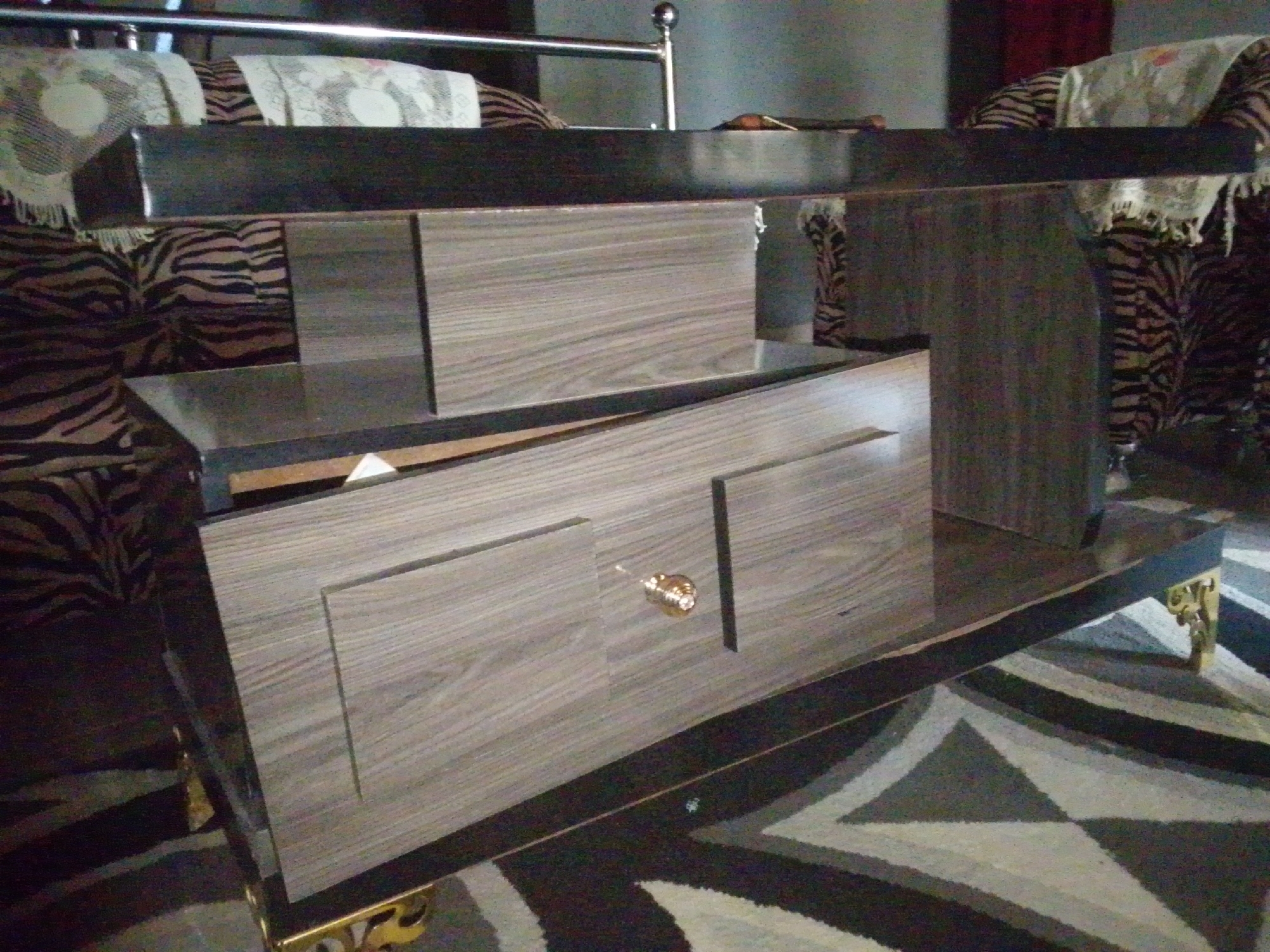 The image shows a close-up of a wooden coffee table with drawers. The table has a dark wood top and lighter wood drawer fronts with a gold knob. It is placed on a patterned rug with geometric designs. In the background, a sofa with a zebra print upholstery and decorative throws can be seen, along with what appears to be part of a bed frame. The overall setting suggests an indoor environment, possibly a living room. The location is identified as Jalingo, Nigeria.