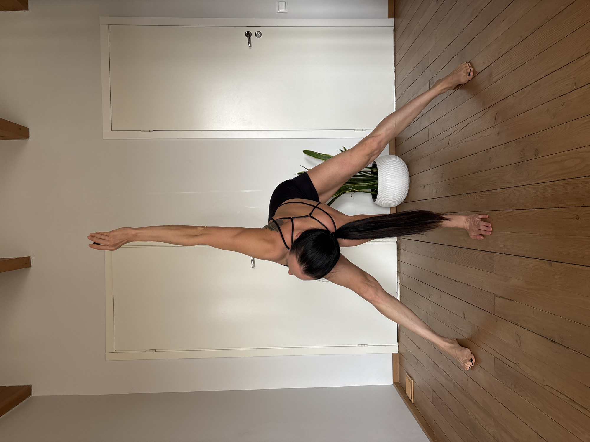 This media file, captured in Chaikovsky, Russia, displays an adult female demonstrating an advanced acrobatic or yoga pose indoors. The image is rotated 90 degrees clockwise, showing the woman horizontally across the frame. She has long dark hair tied in a ponytail and is dressed in black athletic shorts and a black sports bra with intricate back straps. The woman is performing a challenging balance, with her left leg and left arm extended outwards to one side, and her right arm extended in the opposite direction, suggesting a wide standing split or similar flexible posture. Her head is turned to the side, indicating concentration.
The background features light brown wooden plank flooring and neutral-toned walls. Two white door panels are visible on the wall, one larger and one smaller access hatch higher up, along with a small white electrical outlet. A white, textured pot holding a green leafy plant is positioned near the woman's lower body. The scene is brightly lit, consistent with daytime, with soft shadows. No discernible text is present in the image. The overall impression is of strength, flexibility, and focus within a simple, well-lit domestic or studio environment.