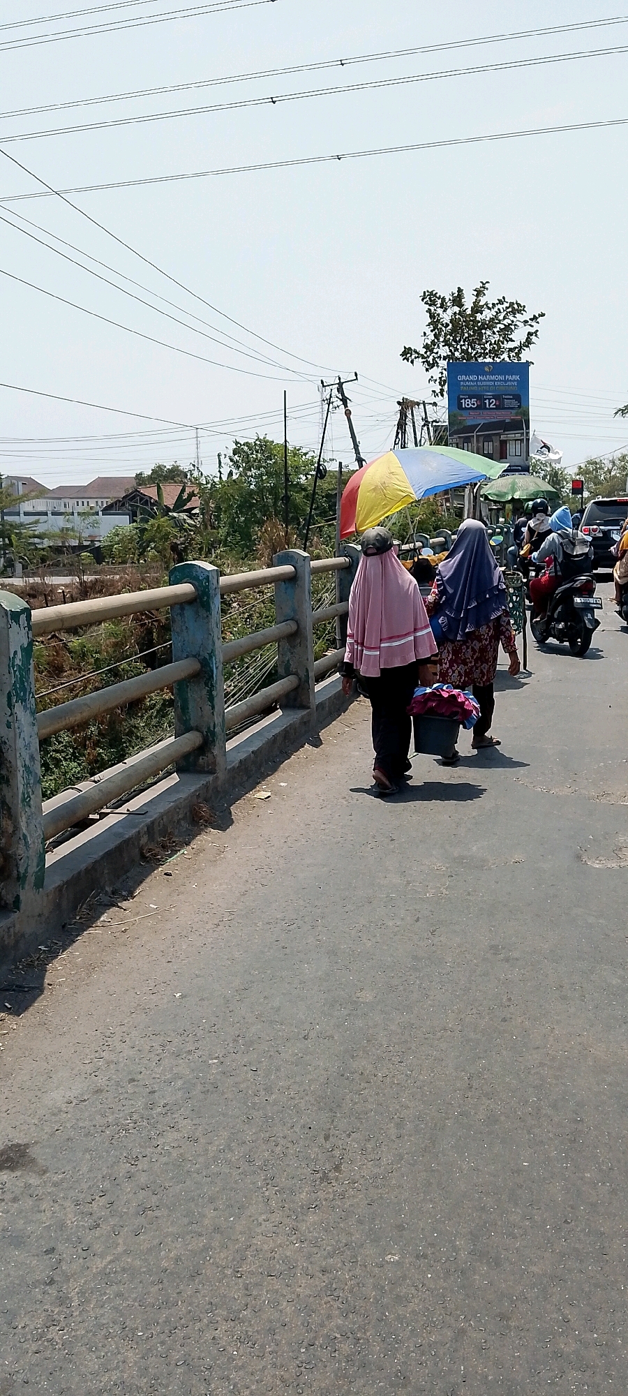 In Srimahi, Indonesia, on a sunny day, two women are walking on a bridge. The woman on the left is wearing a pink headscarf and a dark outfit, carrying a bucket filled with colorful laundry. The woman on the right wears a blue headscarf and a patterned dress, and they are both walking away from the viewer. An open multicolored umbrella shades them. Behind them, traffic is visible, including a motorcycle with riders and a car. The bridge has a concrete railing with metal bars. In the background, trees, buildings, and utility poles with overhead wires are visible. A billboard for "GRAND HARMONI PARK" is also present. The ground is asphalt, with some debris scattered along the edge of the bridge.