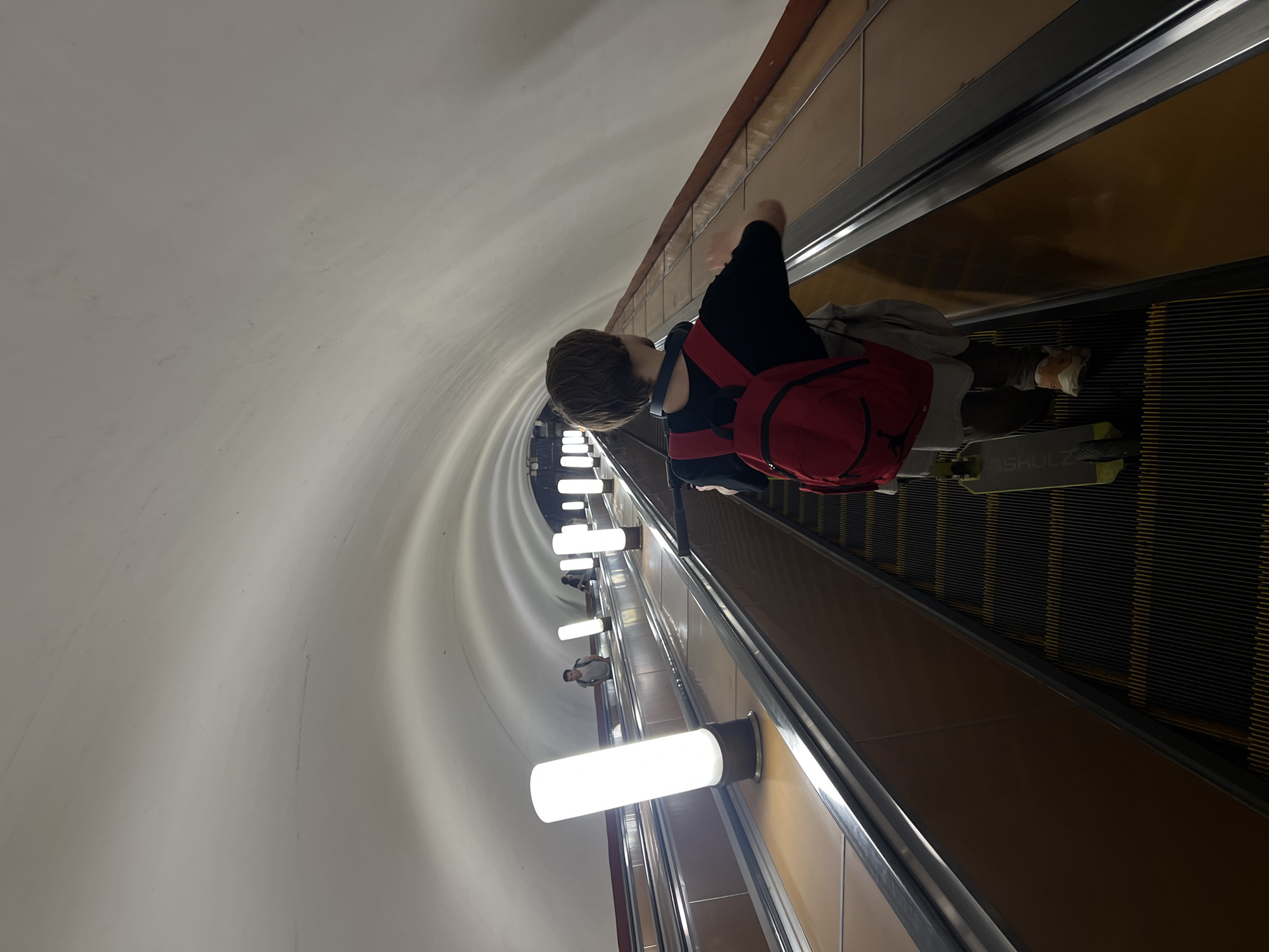 A high-angle photograph shows a person with short brown hair, wearing a black long-sleeve shirt, grey trousers, and a red backpack, riding down an escalator. They are standing near the end of the escalator, and the brand name "SHOLZ" is visible on a black object. The environment consists of a long, curved tunnel with rounded walls, illuminated by a series of tube-shaped lights mounted on the wall alongside the escalator. The escalator itself has yellow-orange walls and black steps. The photograph was taken in the Moscow Metro.