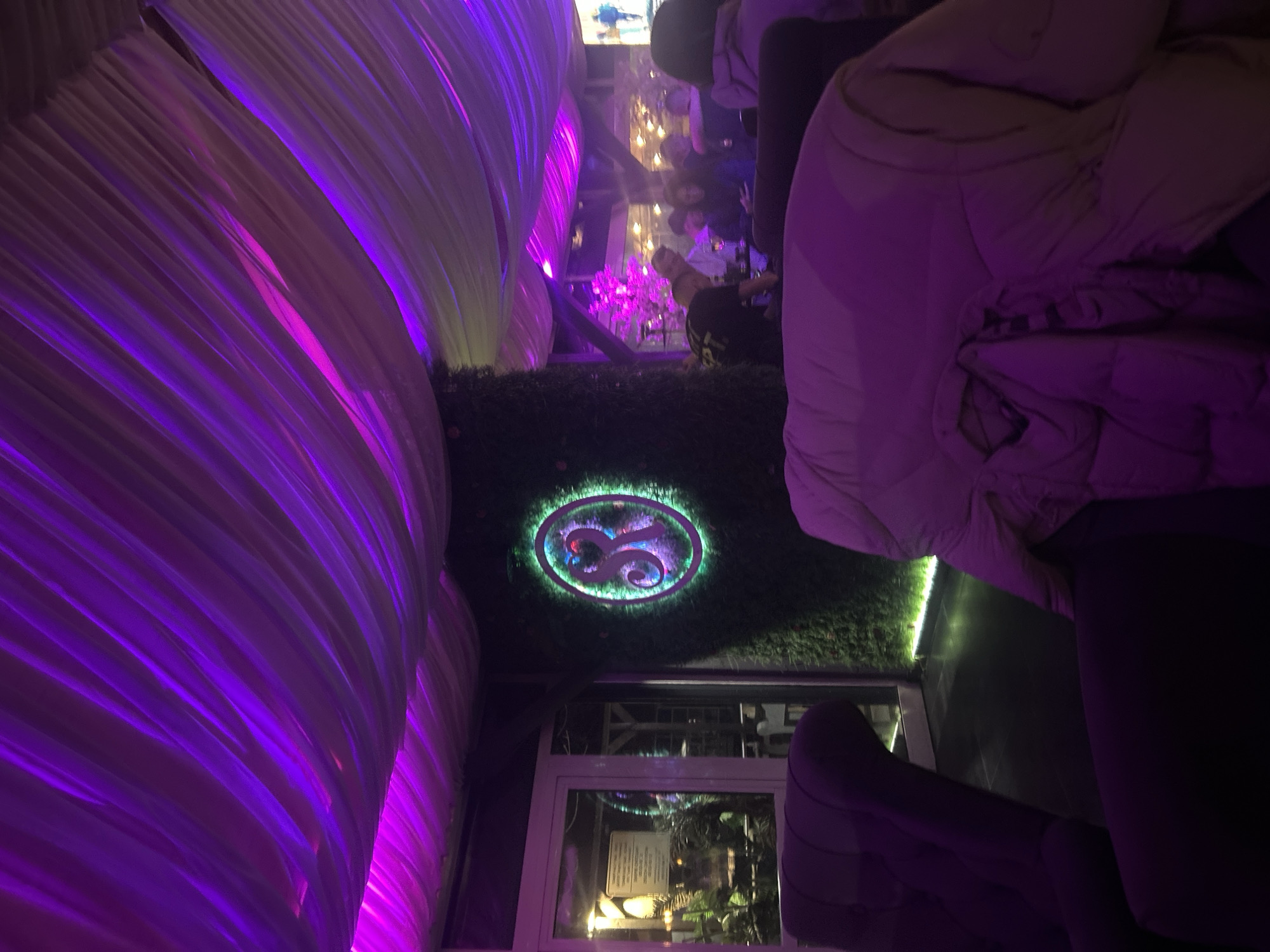 Indoor scene. A white tent-like canopy draped with purple lighting is seen to the left. In the center is a green wall with a circular logo, which is illuminated with green and red lights. Below that is a window or doorway. Occupants are seated on the right, obscured. In the background are additional people. Location details (Zlatograd, Bulgaria) are based on provided context, not intrinsic to the image.