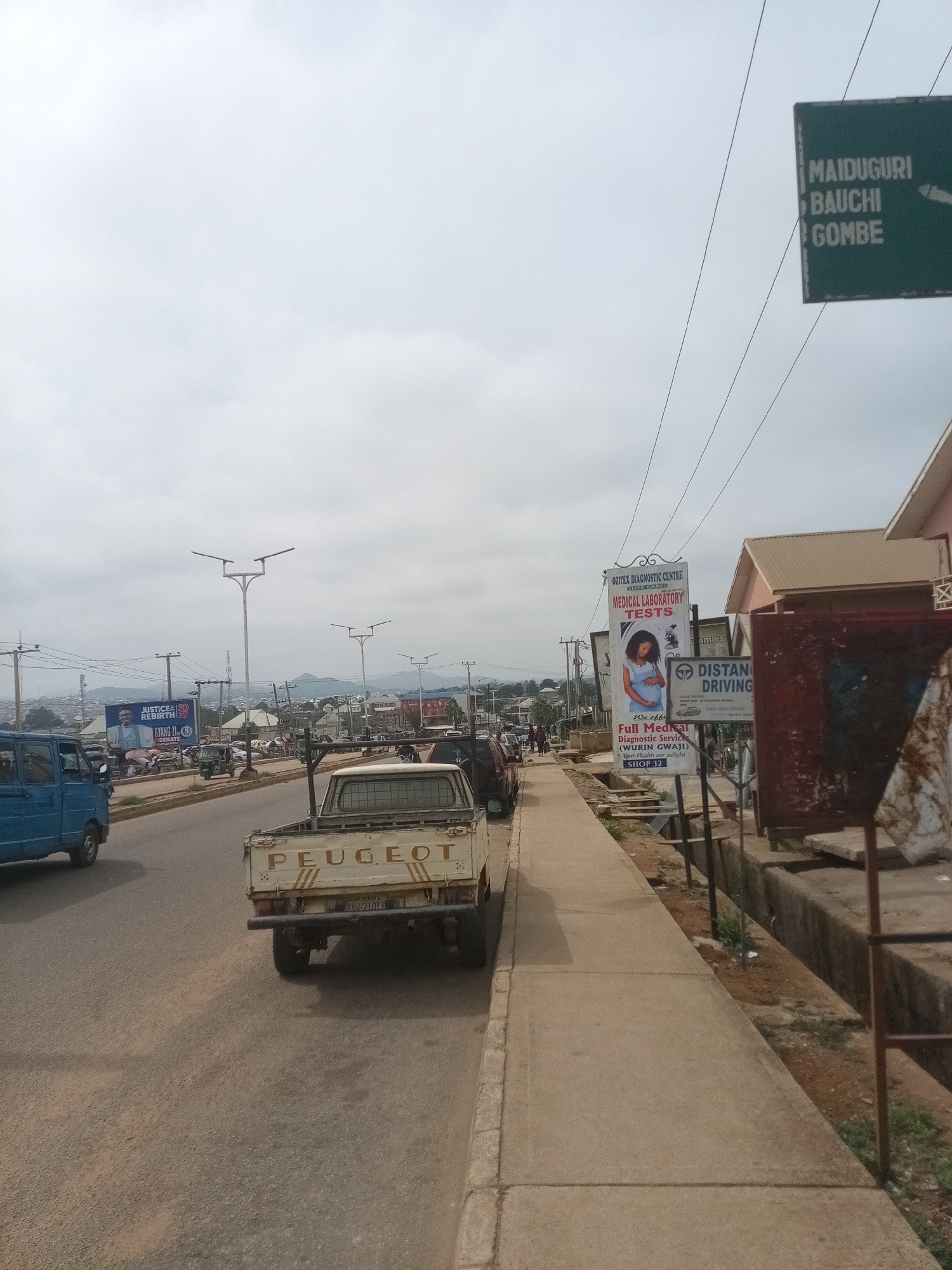 The image depicts a street scene in Jos, Nigeria, characterized by a cloudy sky and a general atmosphere of a bustling urban environment. The street is paved and appears to be a main thoroughfare, with a sidewalk on the right side. Several vehicles are visible, including a blue van on the left, a white Peugeot pickup truck in the foreground, and other cars and a tricycle in the background, suggesting moderate traffic.
Along the sidewalk, multiple billboards and signs are displayed. A prominent green sign on the right indicates directions to "MAIDUGURI," "BAUCHI," and "GOMBE," implying these are major cities accessible from this location. Another billboard advertises "OZITEX DIAGNOSTIC CENTRE" and "MEDICAL LABORATORY TESTS," featuring an image of a pregnant woman. Next to it, a sign for "DISTANT DRIVING" is visible. Further down the street, a large political billboard featuring a man in a suit with the text "JUSTICE 2023 REBIRTH" and "GYANG | SENATE" is also present.
A few individuals are seen walking on the sidewalk and near the vehicles in the background, but their activities are not clearly discernible. The environment includes a mix of commercial establishments, some with visible structures and awnings, alongside utility poles and power lines crisscrossing the sky. The landscape in the distance suggests a hilly terrain surrounding the city. The overall impression is that of a typical commercial street in a Nigerian city.