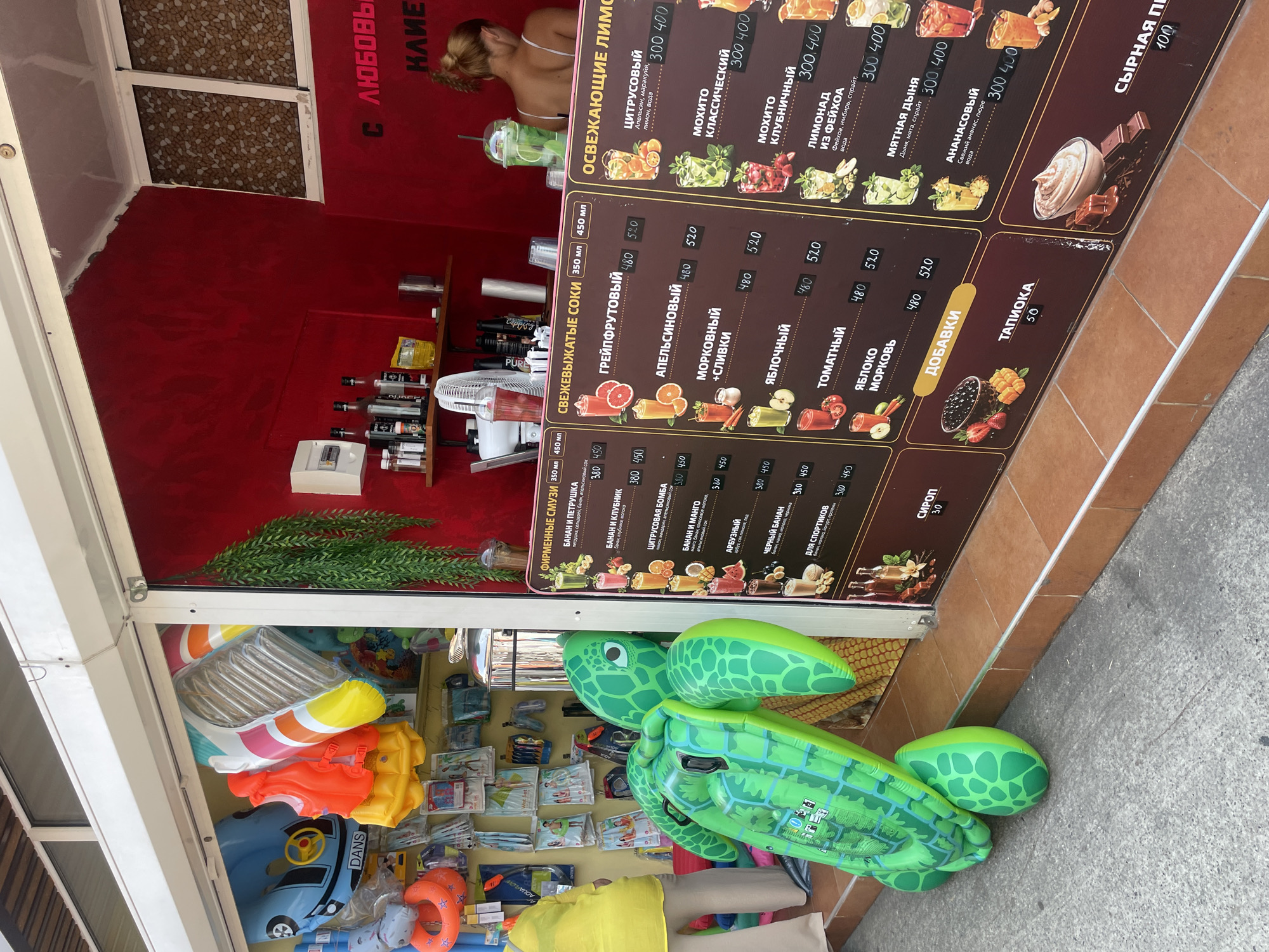 The image depicts a vibrant street-side food and drink stand, likely located in Sochi, Russia, given the Cyrillic text on the menu boards. The stand is characterized by a bright red backdrop and an assortment of colorful inflatable beach toys, including a large green turtle float dominating the foreground. To the left, beach towels, life vests, and other aquatic accessories are displayed. The main menu boards are prominently featured, advertising a variety of fresh juices, smoothies, and lemonade in different sizes and prices.
The menu lists items such as "Фирменные смузи" (Signature Smoothies) with options like "Банан и петрушка" (Banana and Parsley), "Банан и клубника" (Banana and Strawberry), "Цитрусовая бомба" (Citrus Bomb), "Банан и манго" (Banana and Mango), "Яблочный" (Apple), "Черный банан" (Black Banana), and "Для спортсменов" (For Athletes). There are also sections for "Свежевыжатые соки" (Fresh Juices) with choices like "Грейпфрутовый" (Grapefruit), "Апельсиновый" (Orange), "Морковный с сливками" (Carrot with cream), "Томатный" (Tomato), and "Яблоко морковь" (Apple carrot). Another section advertises "Освежающие лимонады" (Refreshing Lemonades) including "Цитрусовый" (Citrus), "Мохито классический" (Classic Mojito), "Мохито клубничный" (Strawberry Mojito), and "Лимонад из фейхоа" (Feijoa Lemonade). A separate "Добавки" (Add-ons) section is visible, along with "Тапиока" (Tapioca) and "Сырная пл" (Cheese pl), the latter likely referring to a cheesecake. The prices are indicated in rubles.
In the background, a woman with her hair tied up is seen working behind the counter, preparing drinks. Behind her, shelves are stocked with bottles, condiments, and other supplies. A small white fan is positioned near the counter. The overall scene suggests a busy, tourist-oriented establishment on a warm day, with the focus on refreshing beverages and beach-related merchandise.