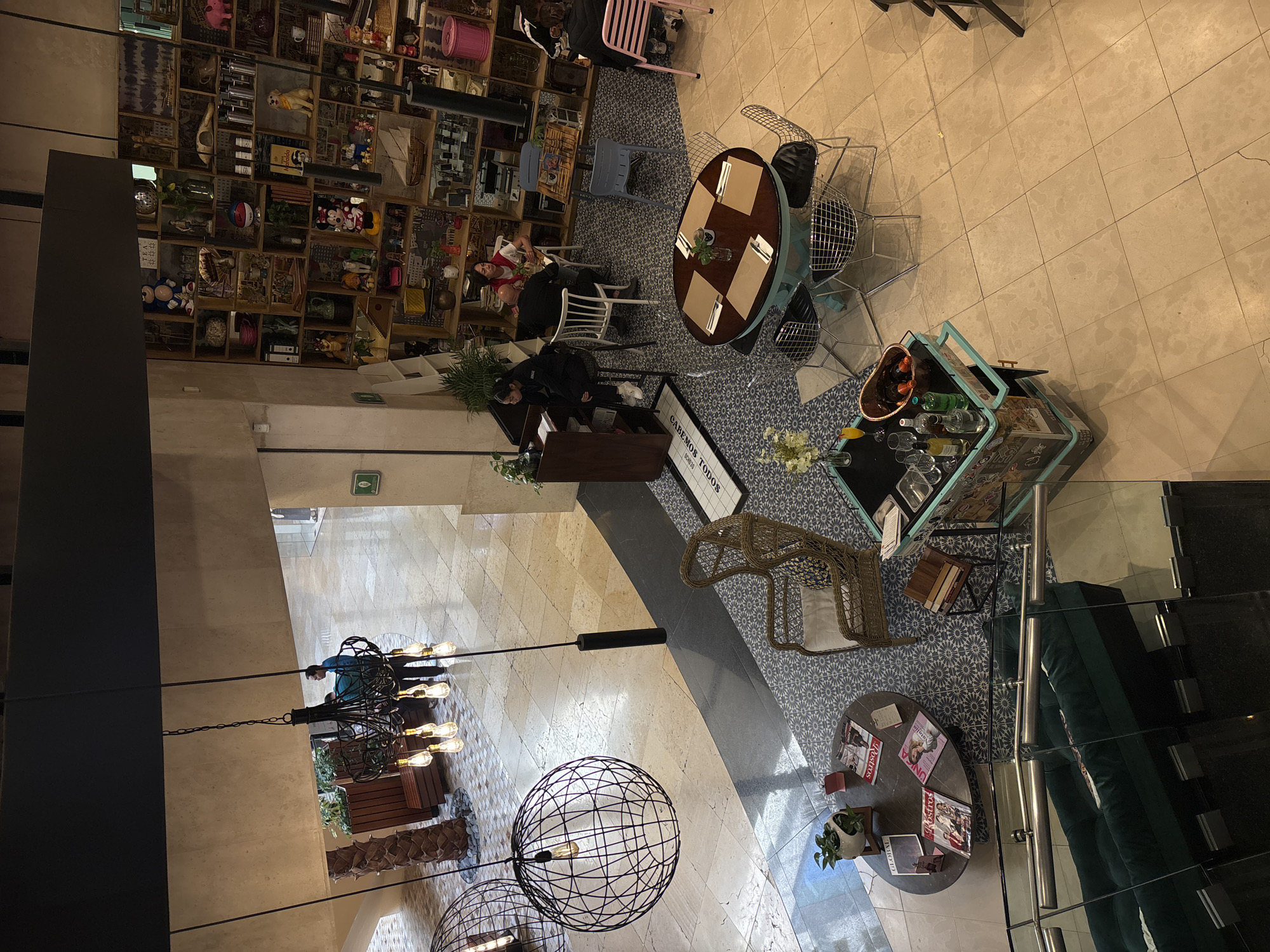 This interior view, likely in Puebla, Mexico, shows a mixed-use space resembling a restaurant or cafe situated within a building featuring a display wall filled with various objects. A couple is seated at a table adjacent to the service counter. The space incorporates various seating arrangements, including tables and chairs, along with decorative elements like lighting fixtures, a bar cart with bottles, and plants. The flooring consists of a mix of tile patterns. There is a glass barrier that obscures some items.