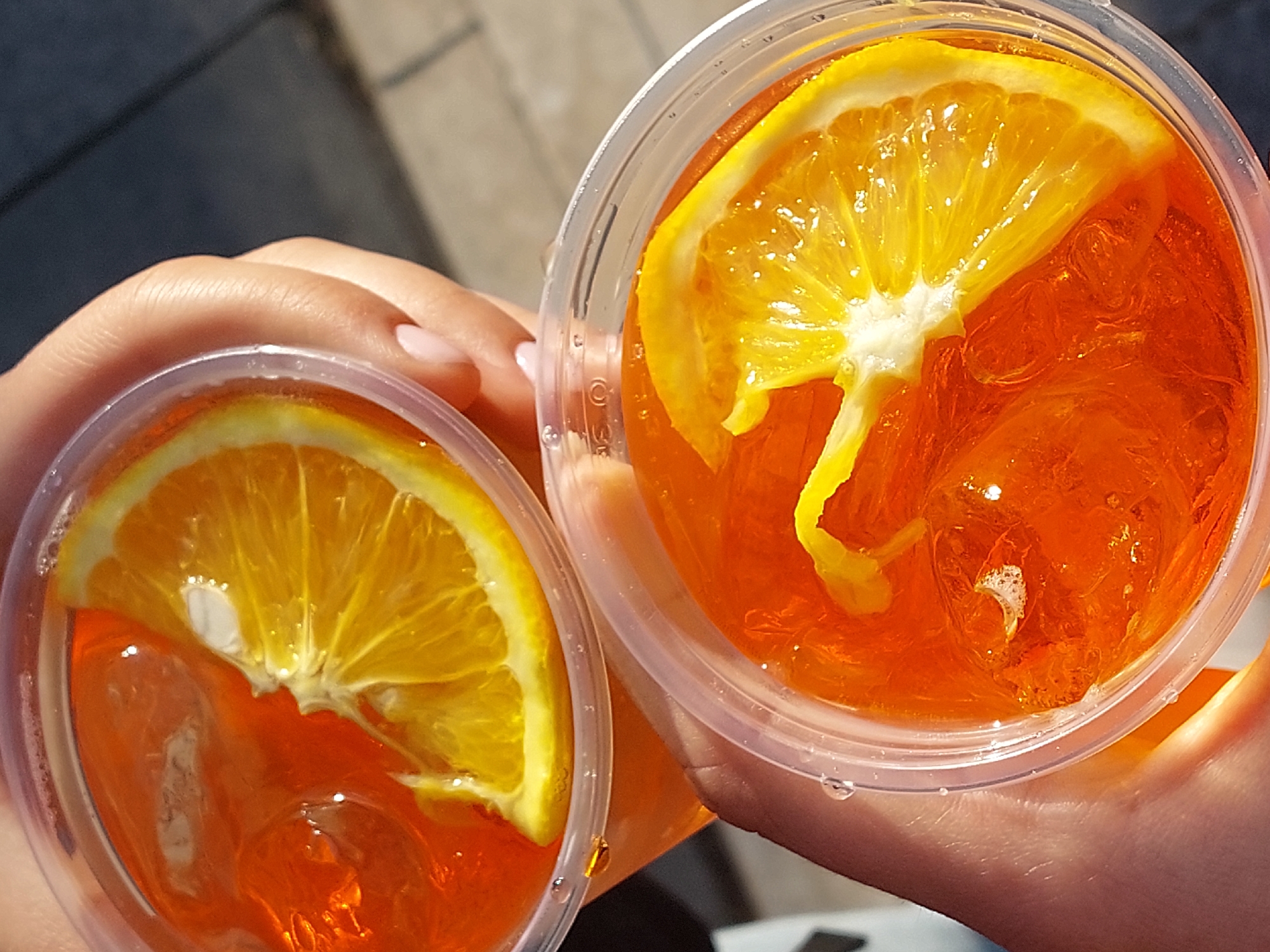 A close-up shot depicts two clear plastic cups filled with an orange liquid, presumably a beverage, likely taken outdoors in the city of Unknown, Unknown. Each cup contains ice and a slice of orange. One cup has a more vertically oriented orange slice, while the other has a more horizontal slice. A hand holds both cups, suggesting the photo was taken by someone capturing the drinks. The cups appear to be freshly made, and the overall setting implies a casual, possibly social, moment. The lighting suggests a sunny day.
