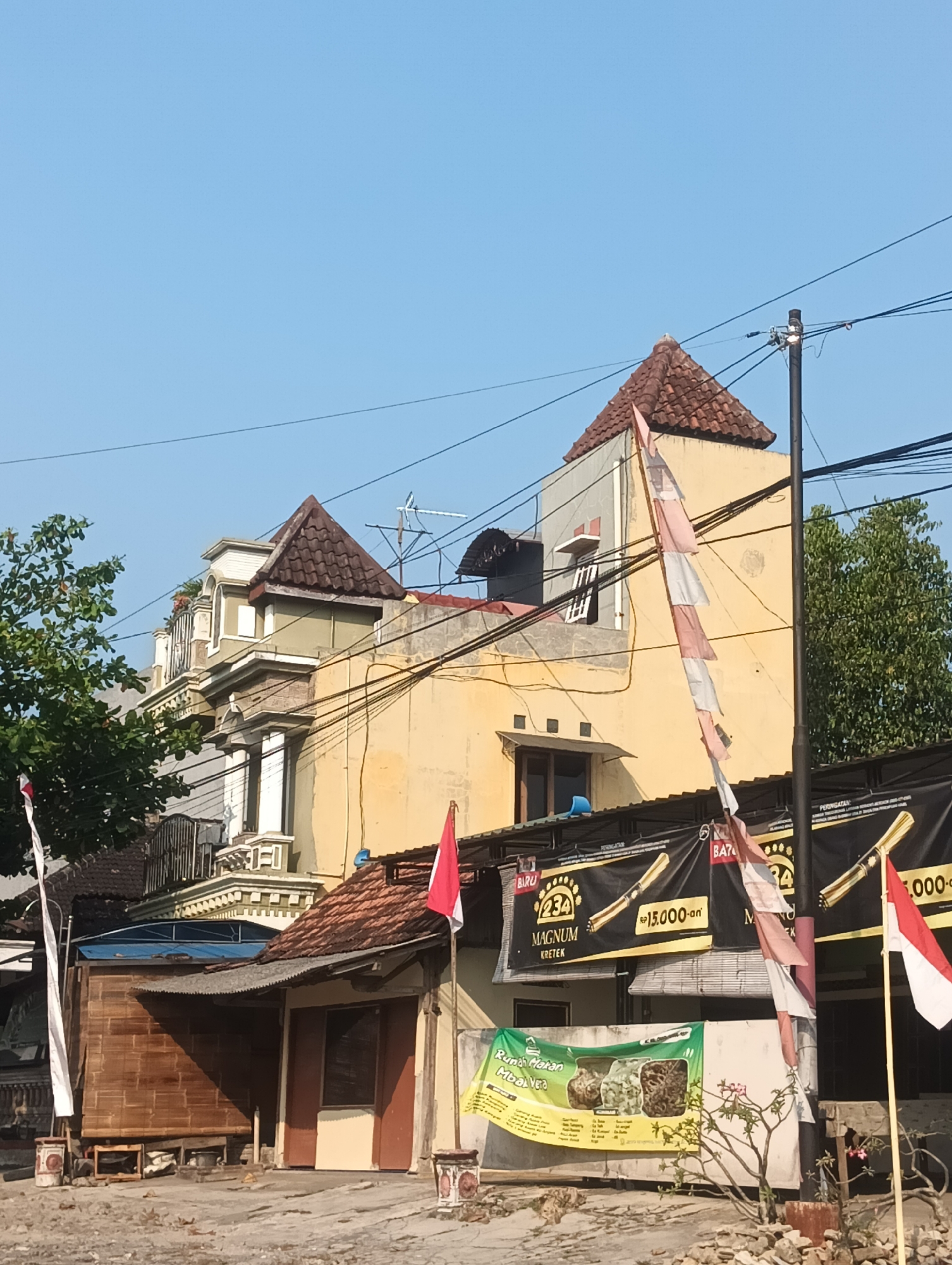 The image depicts a street scene in Palemrejo, Indonesia, characterized by a building with a yellow facade and a multi-tiered roofline. Several red and white flags, typical of Indonesian national symbols, are prominently displayed, some tattered and hanging from poles or attached to buildings. The building's architecture features distinct roof shapes, balconies with ornate railings, and windows of various sizes. Electrical wires crisscross the sky above the scene, supported by a utility pole on the right. In front of the yellow building, a lower structure with a corrugated metal roof is visible, constructed with bamboo walls and a brown door. Banners with black backgrounds and yellow text are hung on the facade of the yellow building, advertising products and possibly an event, one banner featuring an image of a cigarette and the text "MAGNUM KRETEK". Another banner, green with colorful images, is displayed at a lower level. Trees with green foliage are present on the left and right sides of the image, and the ground appears to be a dirt or gravel surface. The sky is clear and blue.