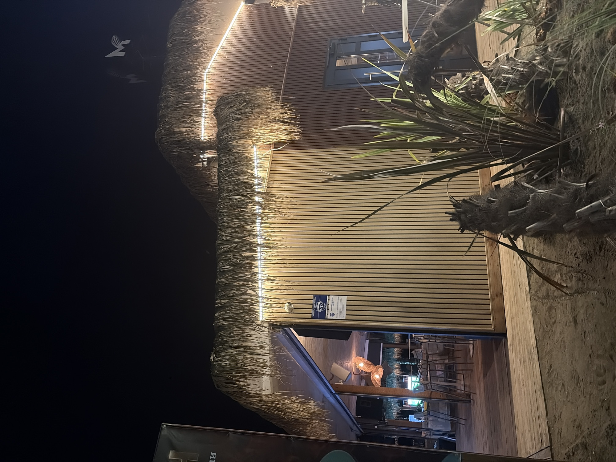 The image shows a beachside establishment at night. The building's exterior is clad in horizontal wooden planks, and a thatched roof overhangs the entrance. LED strip lights illuminate the edge of the roof. A palm tree with long, frond-like leaves is situated to the right of the building. Inside, tables and chairs are visible, suggesting it is a restaurant or bar. The ground is sandy, characteristic of a beach location. There is some visible text on a sign on the building and another sign partially visible at the bottom of the image, though it is mostly obscured. The overall atmosphere is serene and inviting, suggesting a pleasant evening at a seaside venue.