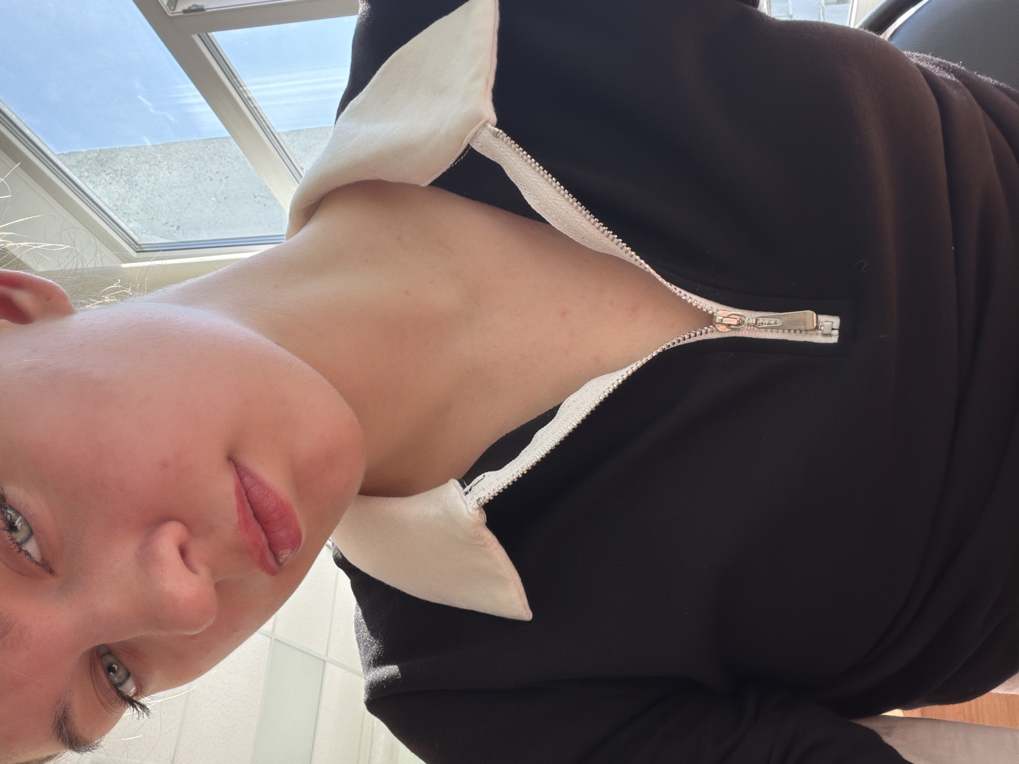 A close-up, slightly angled shot depicts a fair-skinned individual with light green eyes, red lips, and light blonde hair. The person is wearing a black top with a white collar that is partially zipped down, revealing the neck area. The background shows a bright, possibly skylit environment, suggesting an indoor location during daytime. Based on the reflection and light, it appears to be a modern architectural structure, possibly in a city environment. The exact location in Chaikovsky, Russia, cannot be determined based on the visual data.