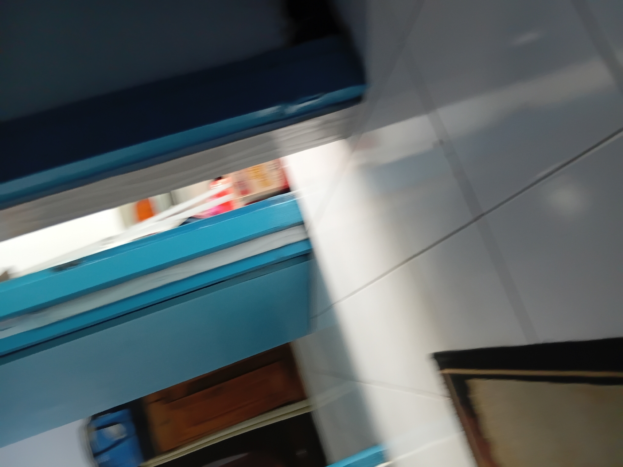 The image shows an interior scene with tiled flooring and doorways. The flooring appears to be light-colored ceramic tile. There are at least two doorways visible, one painted a bright blue and another a darker color. In the background, through the blue doorway, stacked boxes are visible, suggesting storage or a room. To the left, a wooden cabinet or desk is partially visible. The lighting creates distinct shadows and reflections on the floor. The overall impression is of a hallway or room within a building. The scene is likely located in Tanggul Wetan, Indonesia, based on the provided information.