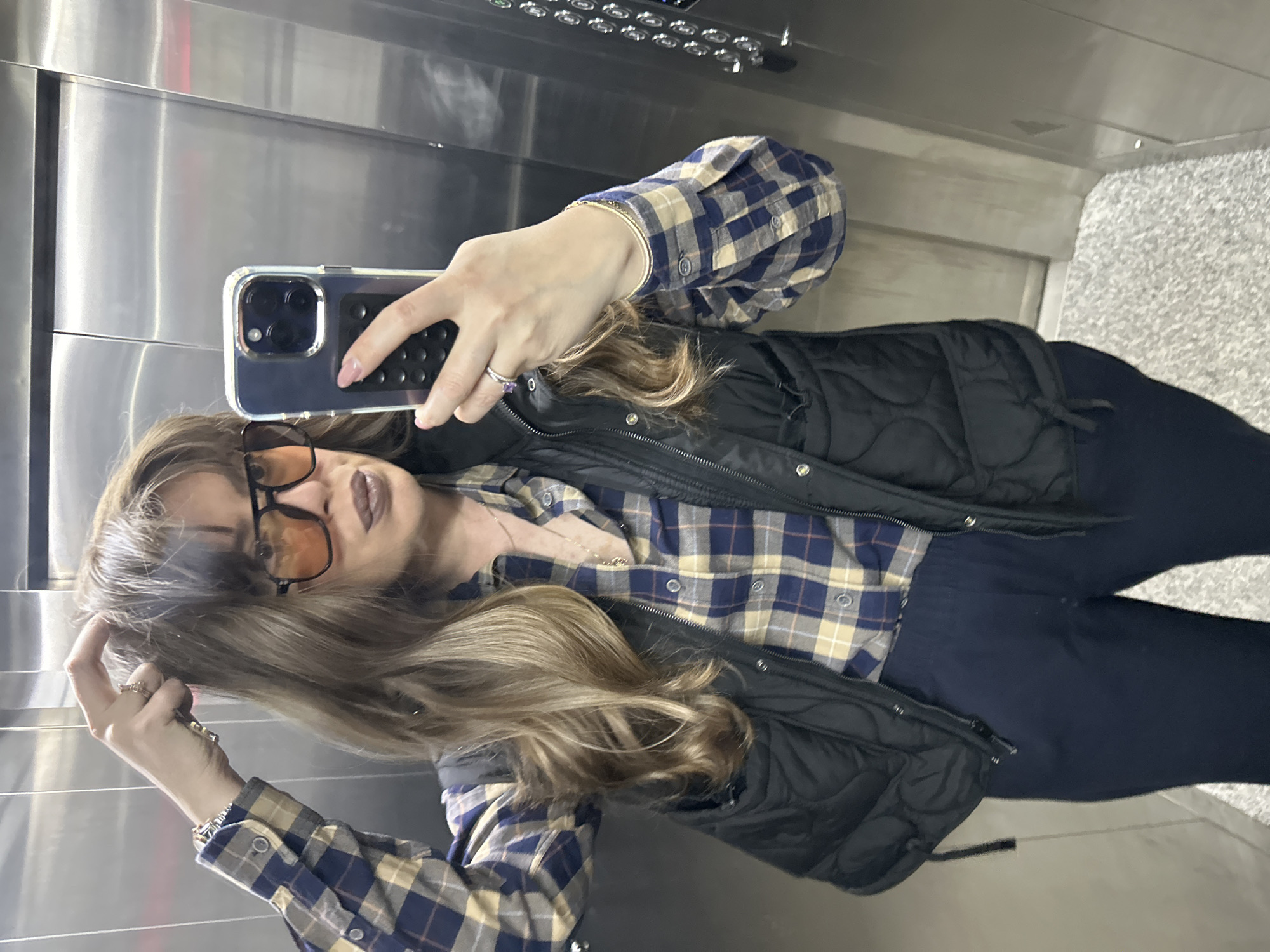 A woman is taking a selfie in an elevator with a phone in a clear case. She is wearing glasses with orange lenses, a blue and beige plaid shirt, and a black quilted vest. She is standing in an elevator with stainless steel walls. A control panel with multiple buttons is visible above her. The floor is gray and speckled. The location is likely in Tbilisi, Georgia.
