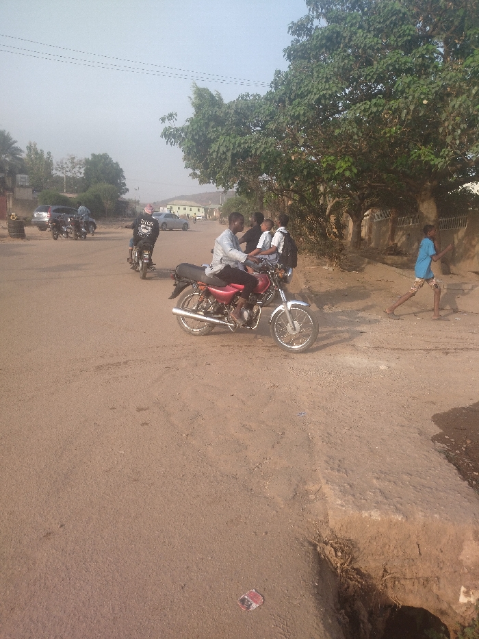 The scene takes place on a dirt road in New Karu, Nigeria, at the coordinates 9 latitude, 7.557 longitude. A man on a red motorcycle is stopped in the middle of the road. There are three other men standing near the motorcycle, two are in white shirts and one is wearing a black shirt. One of the men in a white shirt has a backpack on. In the background, another man in a black shirt is riding a black motorcycle. There are other cars and motorcycles parked on the side of the road. There is a large tree on the right side of the road. A young boy is walking away from the motorcycle towards the tree. There is a large ditch to the right of the road that appears to have eroded away. The scene is sunny and the sky is clear.