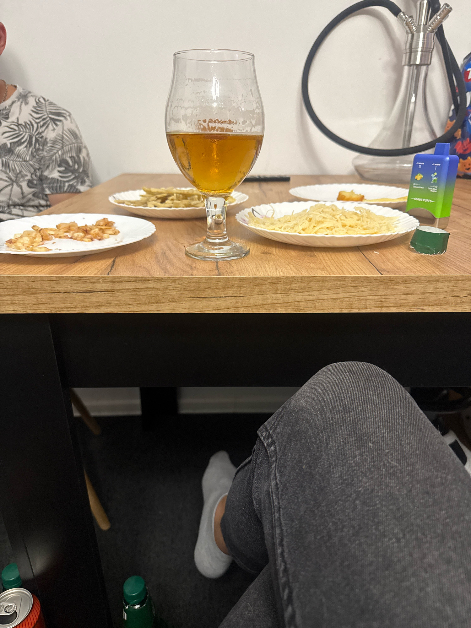 In an indoor setting, likely a dining area, a table is laden with food and drinks. A person's legs, clad in dark jeans and grey socks, are visible in the foreground. On the table, a glass of amber liquid, possibly beer, stands prominently. Beside it, plates hold snacks, including what appears to be shredded cheese and fried pieces of food. A hookah pipe is visible in the background, suggesting a relaxed social gathering. A bright blue vape device rests on the table. The overall impression is one of casual dining and leisure. The specific location in Gorzów, Poland, cannot be determined from the image alone.