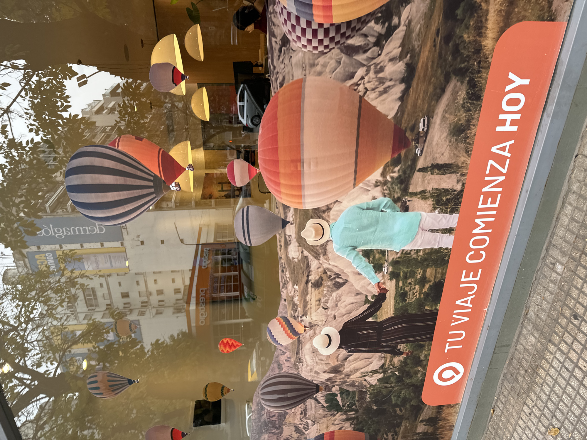 This photo depicts a storefront display in Buenos Aires, Argentina, featuring an advertisement for travel, superimposed with reflections of the street outside. The advertisement showcases a scenic landscape with hot air balloons and images of two people holding hands. The text "TU VIAJE COMIENZA HOY" is prominently displayed. The reflections include the street and buildings across the street.