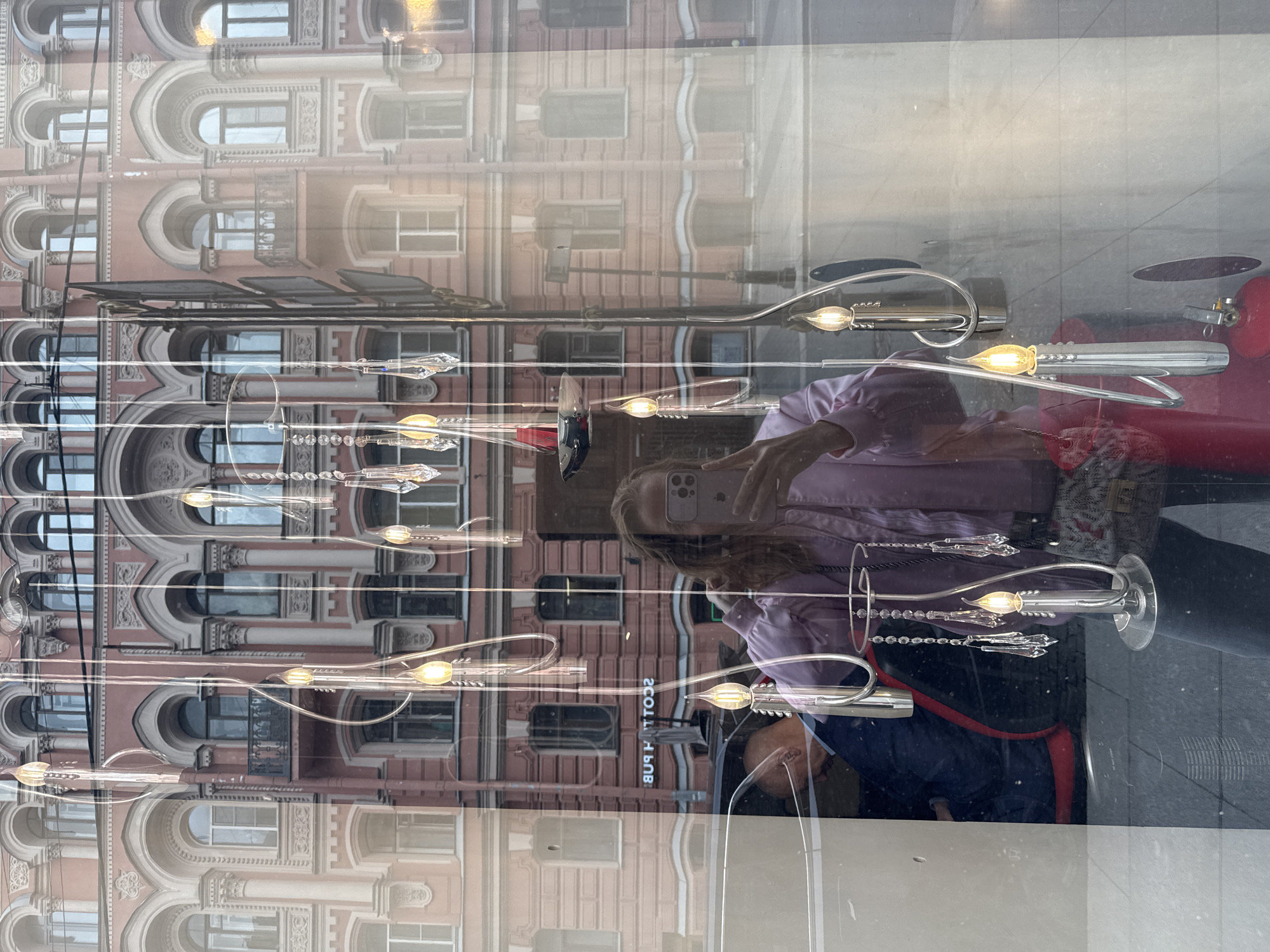 The image shows a reflection in a window in Saint Petersburg, Russia. The reflection includes a pink building with arched windows, likely a historic structure characteristic of the city's architecture. Inside the window are suspended light fixtures with bulbs. A person is reflected in the window, holding a mobile phone and taking a picture. Below the person is a reflection of a person wearing a jacket.