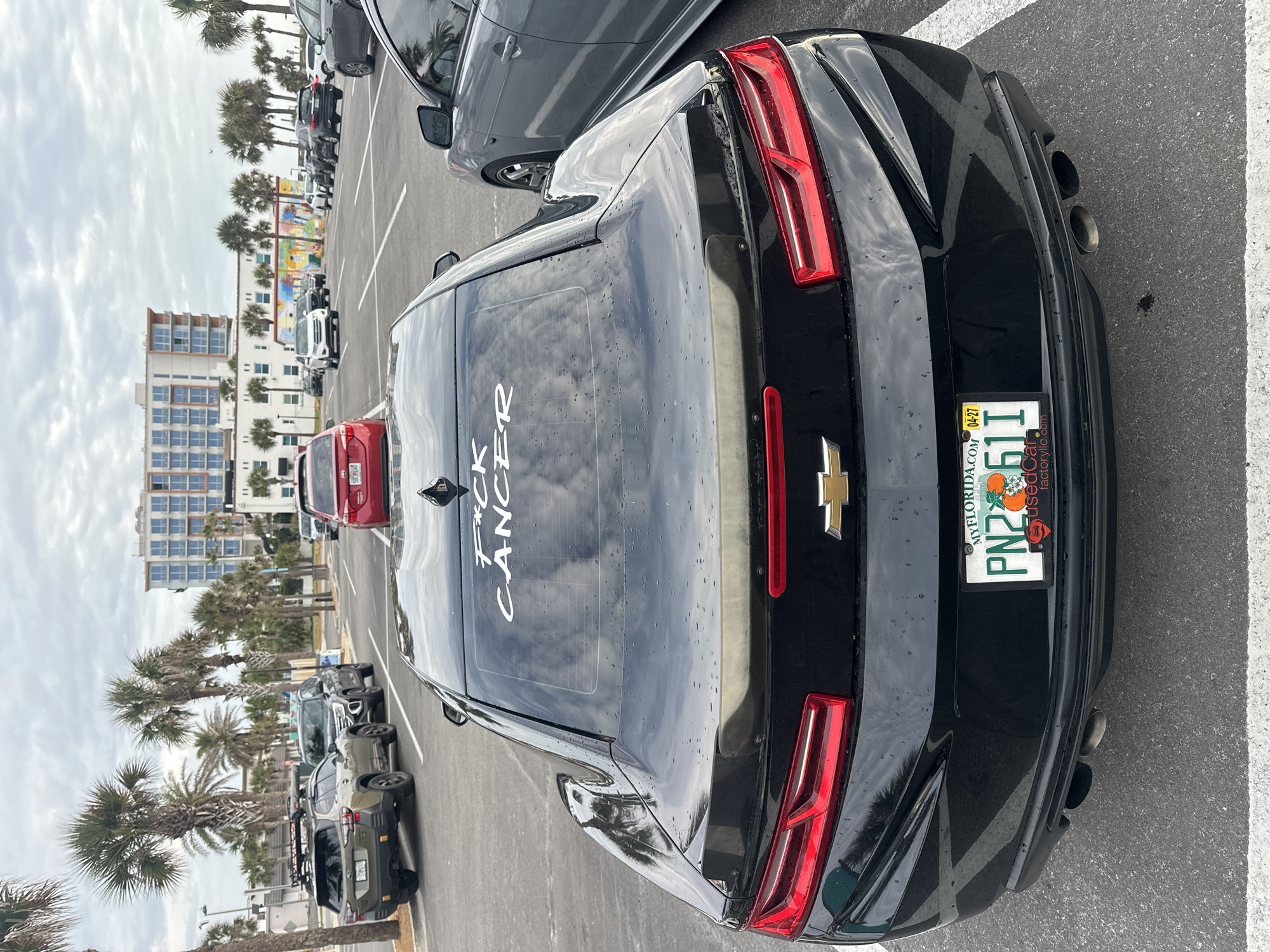 The image shows a black Chevrolet Camaro parked in a parking lot in Jacksonville Beach, Florida, United States of America. The car has a Florida license plate with the number PN2 61T. The phrase "F*ck Cancer" is written in white on the rear window of the vehicle. In the background, there are palm trees, buildings, a colorful mural, and other parked cars. The day appears overcast.