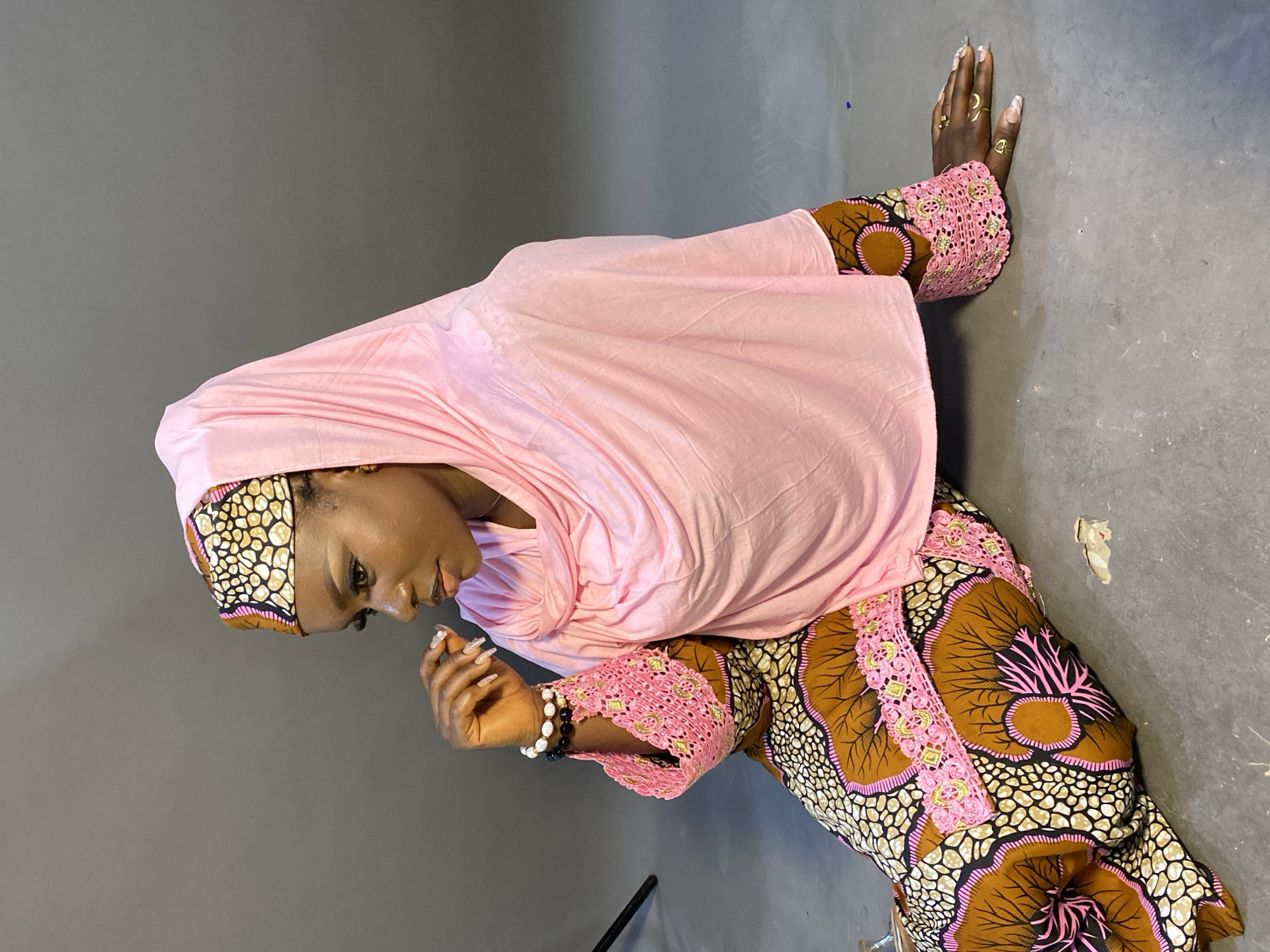 A young woman is depicted kneeling or crouching on a plain, dark grey floor in an indoor setting. She is dressed in a light pink head covering (possibly a hijab or khimar) that drapes over her shoulders and upper body. Her attire also includes a long-sleeved garment made from a vibrant, patterned fabric featuring shades of brown, black, beige, and pink, with intricate, abstract designs. The sleeves and possibly the front of her garment are embellished with pink lace detailing that includes gold accents.
Her head is tilted downwards, and her gaze is directed towards her left hand, which is held up near her chest. On her left wrist, she wears two beaded bracelets, one black and one white. Her right hand rests flat against the grey floor, and rings are visible on her fingers, including one with a crescent moon shape. Her fingernails are long and appear manicured with a light or glittery polish.
The background consists of a plain grey wall, offering a simple and uncluttered backdrop that highlights the subject and her detailed clothing. A small black object, possibly a tripod leg, is visible in the lower left corner of the frame. The woman's pose and downward gaze suggest a moment of contemplation or serenity. No visible text is present in the image.
