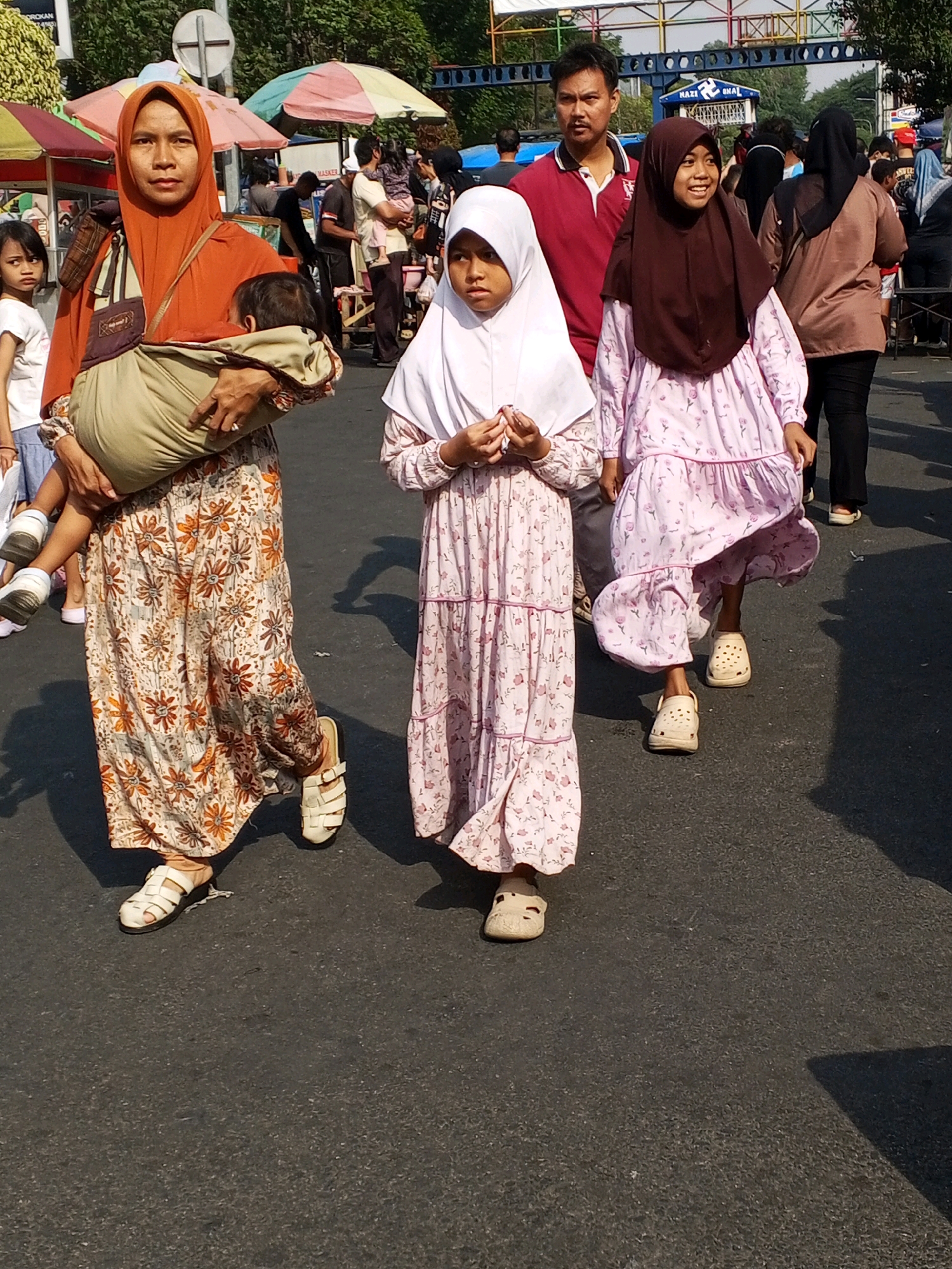 In Serang, Indonesia, a group of people are walking on a paved street. In the foreground, a woman wearing an orange hijab and a long, floral patterned skirt carries a baby in a sling. She is looking forward. To her right, a young girl wearing a white hijab and a pink floral dress walks forward, holding her hands clasped in front of her. Further to the right, another young girl in a brown hijab and a pink floral dress smiles as she walks forward. In the background, more people are visible, along with colorful stalls and umbrellas, suggesting a bustling street market or event. The lighting indicates it is daytime.