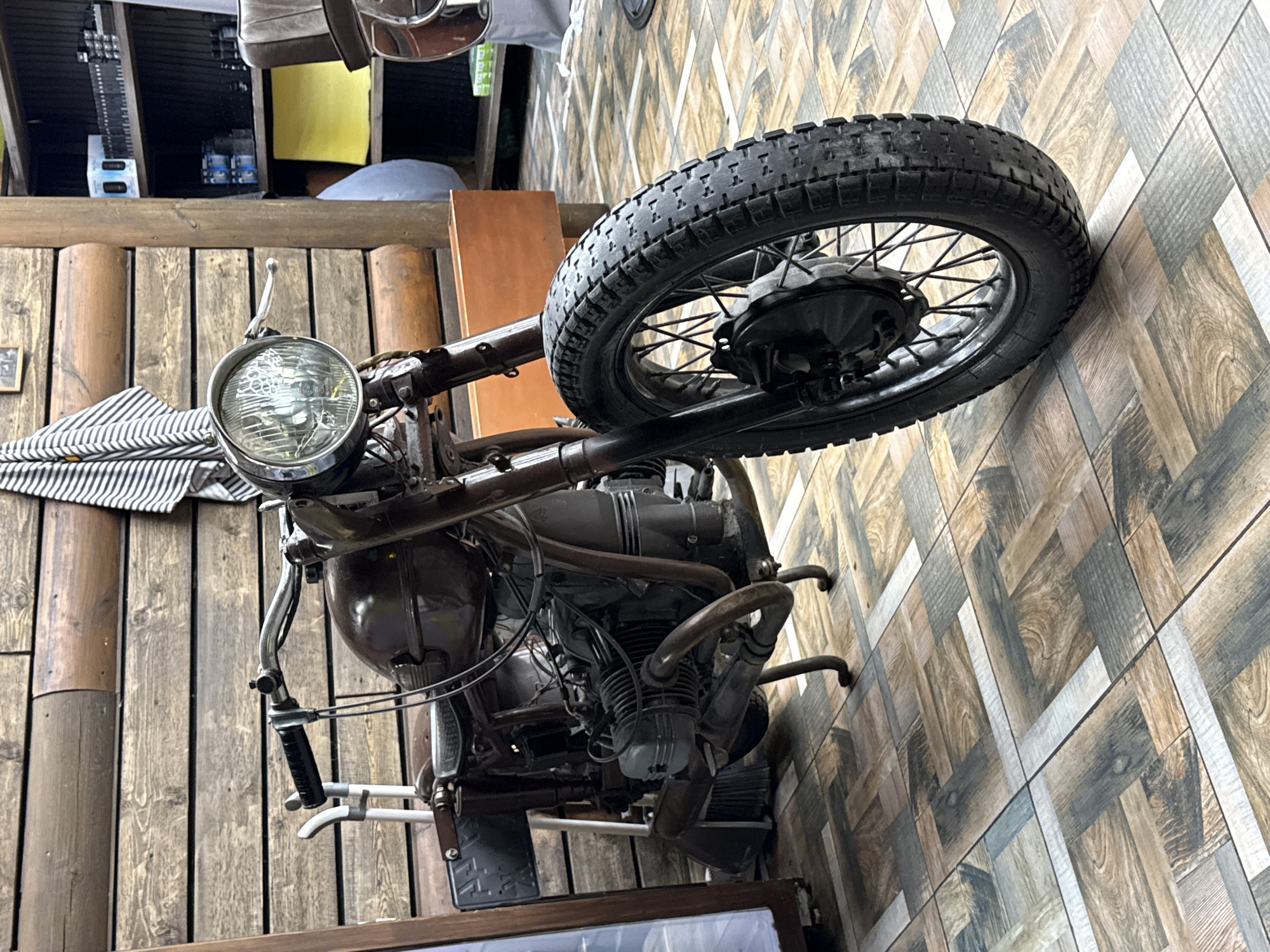 The image captures a vintage or classic motorcycle displayed indoors. The motorcycle is dark brown or bronze, featuring a prominent round headlight, spoked wheels, and knobby tires. Its engine, complete with cylinders and exhaust pipes, is clearly visible, suggesting a well-preserved or restored model. Handlebars with grips and levers are also in view.
The motorcycle is positioned against a background of vertical wooden planks or log-like panels, contributing to a rustic or industrial interior design. To the right, a surface, possibly a floor or another wall, is covered with geometrically patterned tiles that mimic wooden planks in various shades of brown, grey, and white.
In the upper background, shelves hold several objects, including what appear to be bottles or containers. A striped cloth or fabric is draped near the motorcycle's front end. The lighting indicates an indoor setting, likely illuminated by artificial sources. The image is oriented such that the motorcycle appears to be lying on its side, or perhaps it is mounted vertically as a decorative piece. No legible text is visible within the frame. The scene suggests a themed establishment, such as a cafe, bar, or enthusiast's display area.