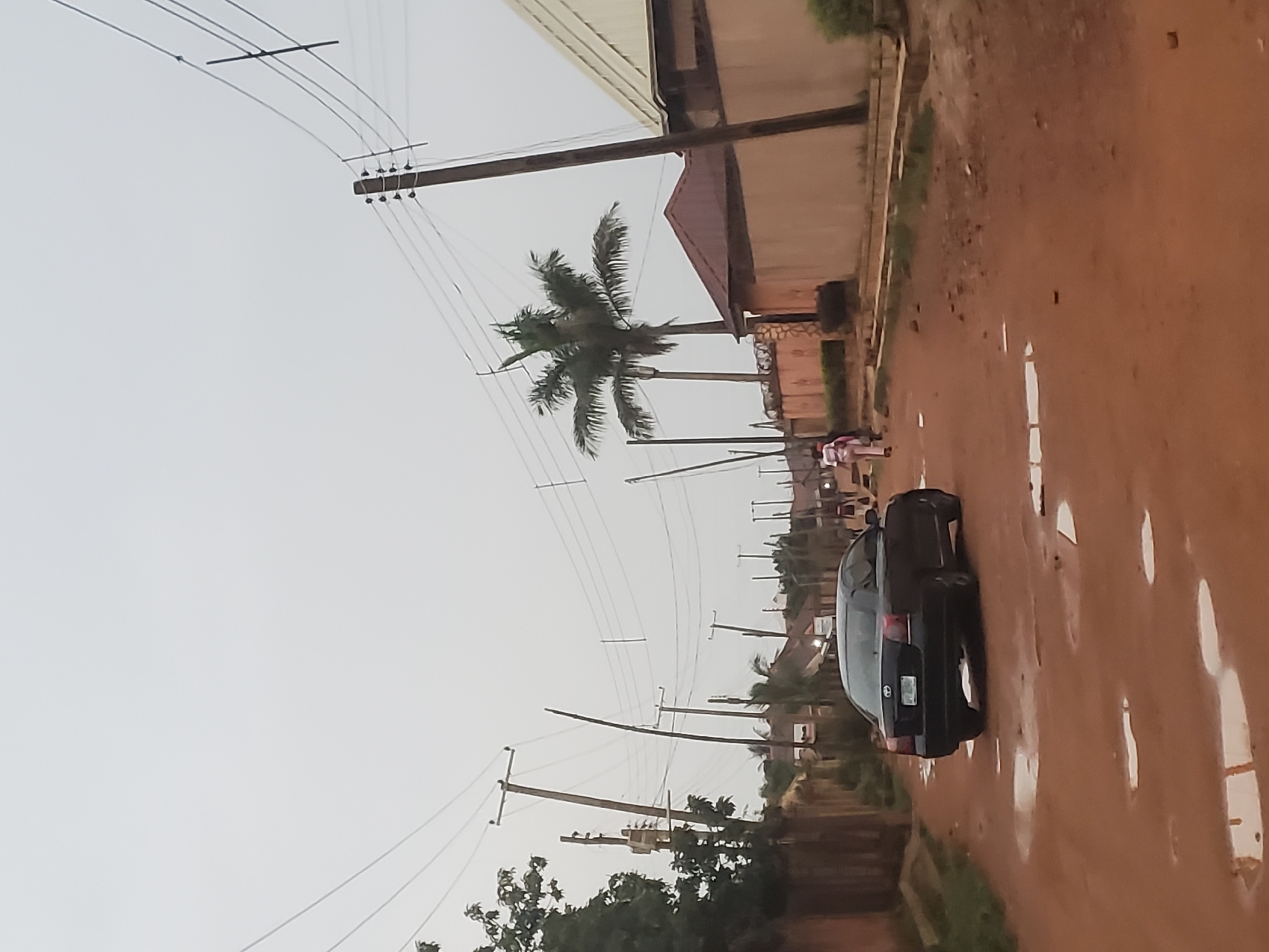 The image depicts a residential street in Barakin Akawo, Nigeria, taken at ground level. The road is unpaved, reddish-brown, and marked with puddles. A dark-colored sedan is parked on the side of the road. Power lines and wooden poles run along the street. A palm tree is visible among the power lines. A building is situated along the street, with a corrugated metal roof. Several people are walking in the distance. The sky is overcast.
