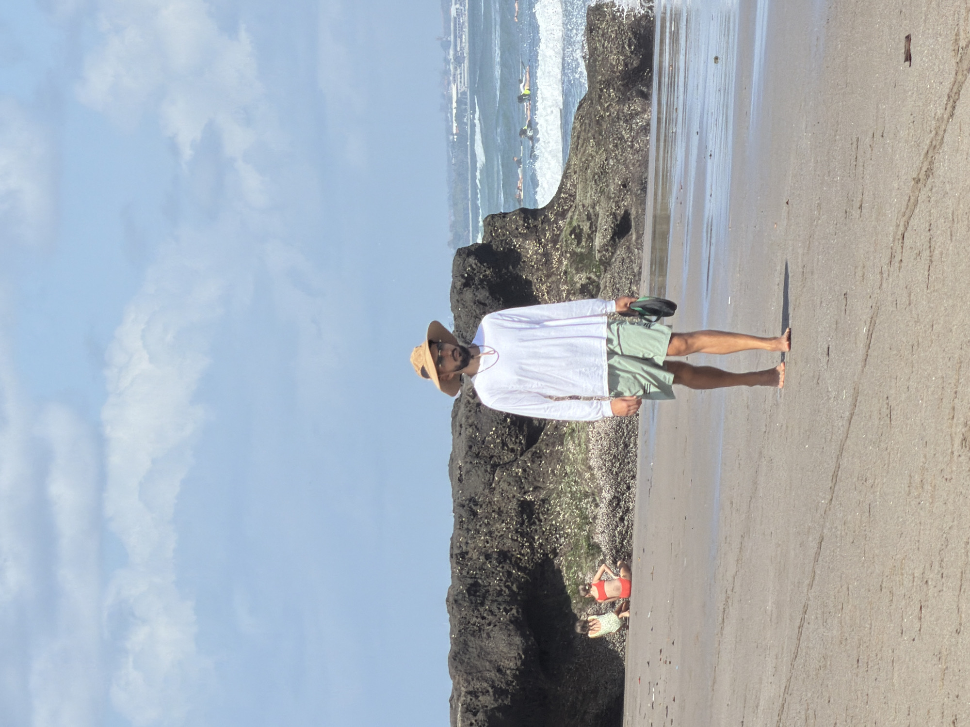 A man wearing a wide-brimmed hat, a long-sleeved white shirt, and light green shorts walks along a sandy beach. He is holding a black flip-flop in his right hand. In the background, two children are sitting on the sand near a rocky outcrop. Further out in the ocean, several people are visible on surfboards. The sky is blue with scattered white clouds. The location is the beach in North Kuta, Indonesia.