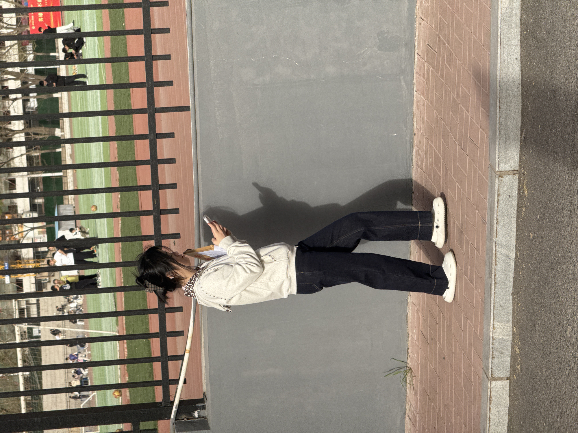 A young woman stands on a brick sidewalk next to a grey concrete wall and a black metal fence, seemingly engrossed in her phone. Behind the fence is a track and field area, with a green field and red track. Several individuals can be seen in the background, some appearing to be students or athletes. The location appears to be an outdoor area near a sports facility in Zhongtun, China.