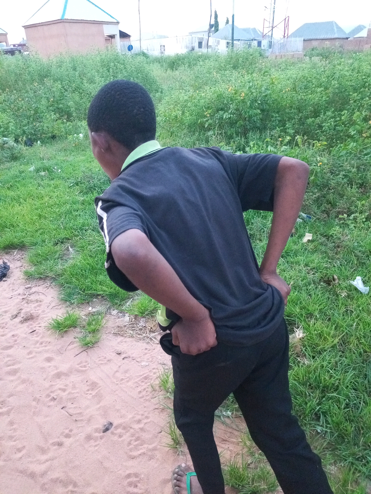 A young boy is standing in an outdoor environment with a sandy area to his left and a grassy area to his right. He is wearing a dark t-shirt with white stripes on the sleeves and black pants. His hands are on his hips. In the background, there are buildings and dense vegetation. This scene is located in Tempure, Nigeria.