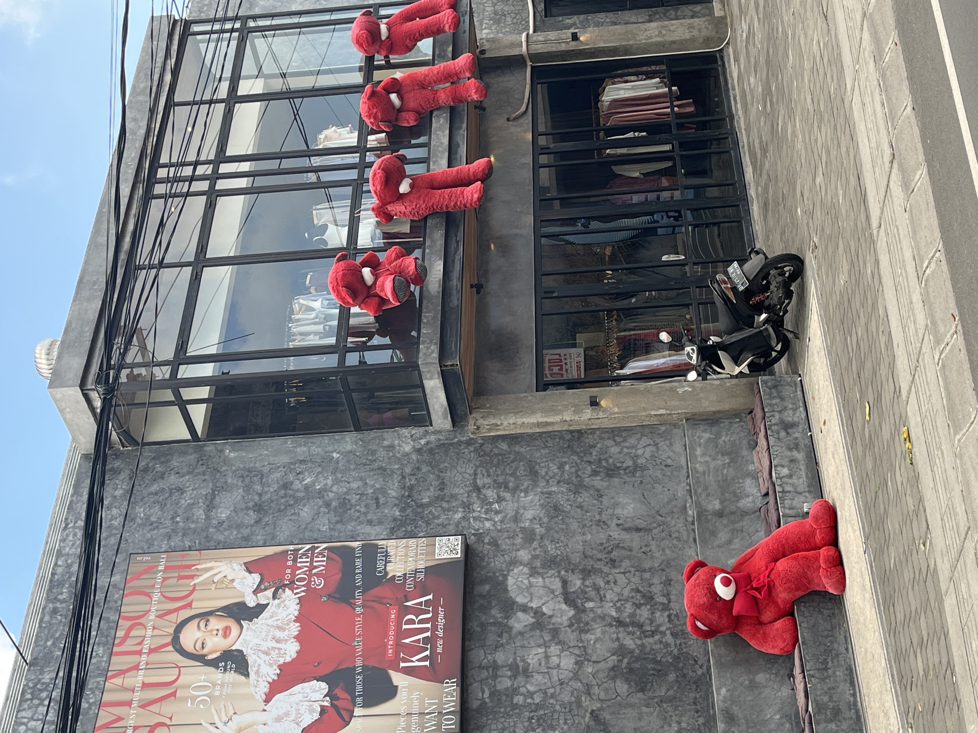 The image captures a street view of a boutique store in North Kuta, Indonesia, with a distinctive facade and an outdoor seating area. The building features a dark, textured concrete wall on the left, dominated by a large, colorful advertisement for "MAISON SAUVAGE" fashion boutique. The advertisement displays a woman in a red jacket and white lace, with text indicating "50+ BRANDS FROM AROUND THE WORLD" and "FOR BOTH WOMEN & MEN." Below the advertisement, a large, plush red teddy bear is seated on a concrete bench, leaning back and facing the street.
The building's upper level has large, dark-framed glass windows, suggesting a storefront or display area. Five more red teddy bears are hanging from the balcony railing of this upper level, their limbs dangling down. Some of these bears appear to be of a smaller size than the one seated outside.
The ground floor of the building has a large glass storefront, revealing clothing racks with various garments. A portion of a dark-colored motorcycle with white accents is parked on the paved sidewalk in front of the store. The sidewalk is made of light-colored square pavers, with a curb separating it from the asphalt street. The environment appears to be a commercial or shopping district, with clear blue sky overhead and some power lines visible against the sky. The overall scene conveys a playful and eye-catching display for the boutique.