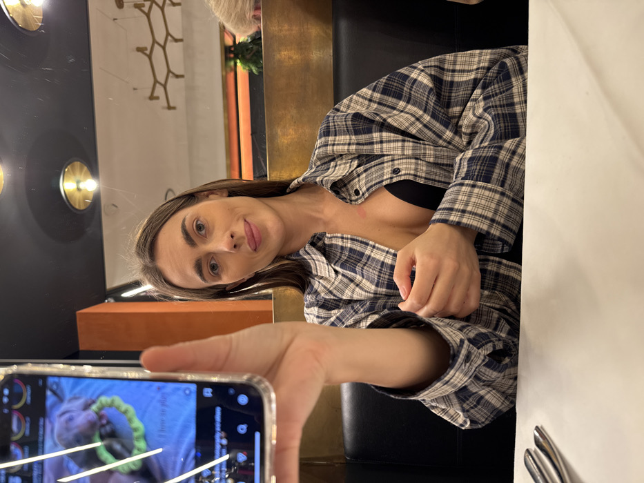 The scene is set indoors, likely within a restaurant or bar in Verona, Italy. A young adult female with long dark hair is seated at a white table, looking directly towards the camera with a slight smile. She wears an unbuttoned blue and white plaid shirt over a black bra, and a faint reddish mark is present on her upper left chest. Her right hand extends forward, holding a smartphone in a clear case. The phone's screen displays an image of a grey puppy playing with a bright green, ring-shaped toy, accompanied by the text "i love to play." Cutlery is partially visible on the left side of the table. The background features black booth seating, a gold-toned partition, an orange vertical panel, and a dark ceiling with circular recessed lights. A mirror reflection in the upper right shows a decorative, geometric chandelier and the partial profile of another individual with light-colored hair.