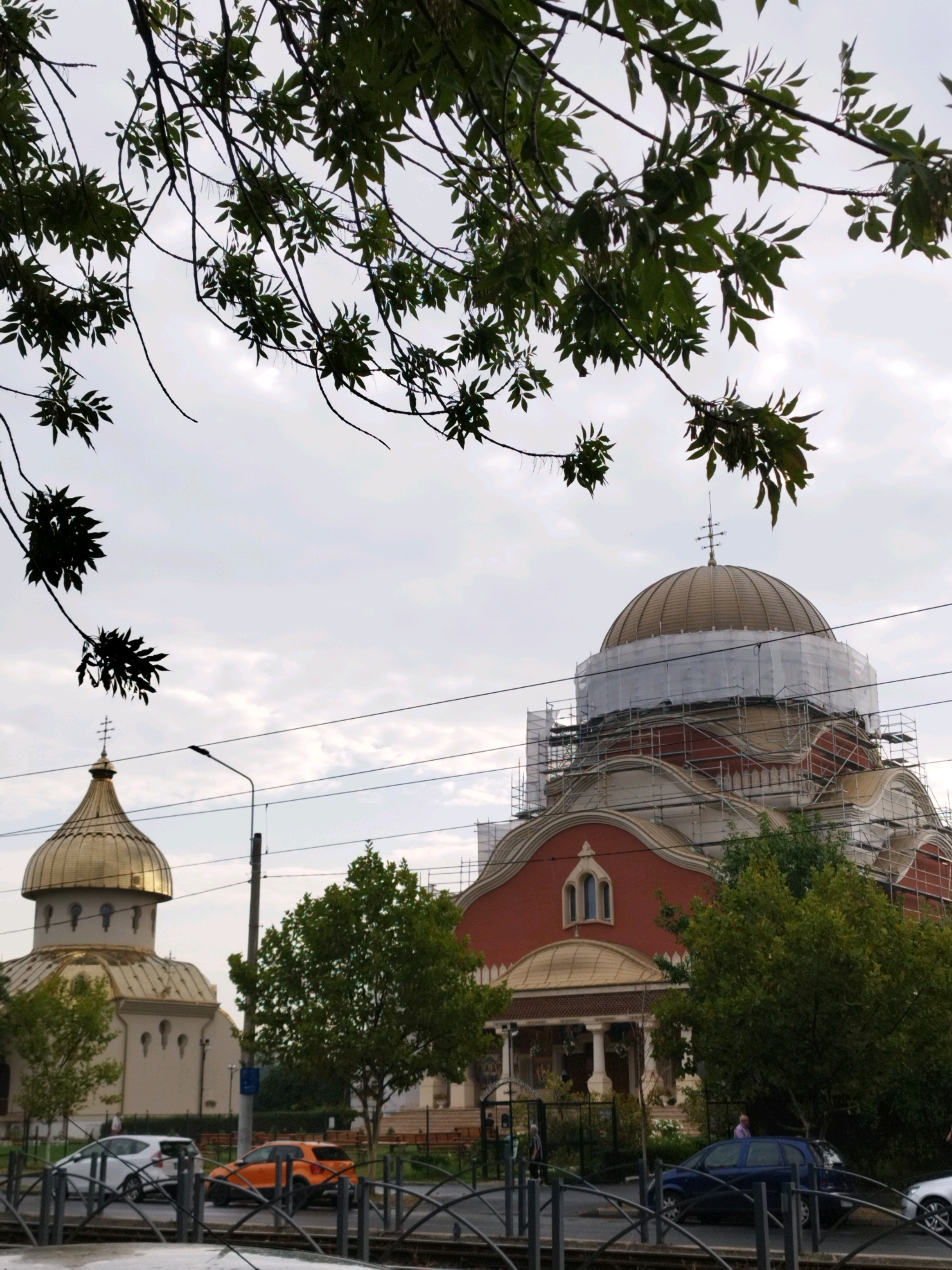 In Bucharest, Romania, an Orthodox church with golden domes is undergoing renovations, as evidenced by the scaffolding and white protective covering around its main dome. The building features red brickwork and arched windows. To the left, a smaller, similarly designed dome of another church is visible. The scene is set against an overcast sky. In the foreground, a street with parked cars, including a white sedan and an orange SUV, runs alongside the church complex. A black metal fence separates the street from a paved area with trees and benches. Several individuals are present, including one near the entrance of the smaller church and another walking on the sidewalk near a blue car. Overhead, branches with green leaves partially obscure the sky and the tops of the church structures. Electrical wires and a lamppost are also visible, indicating an urban environment.