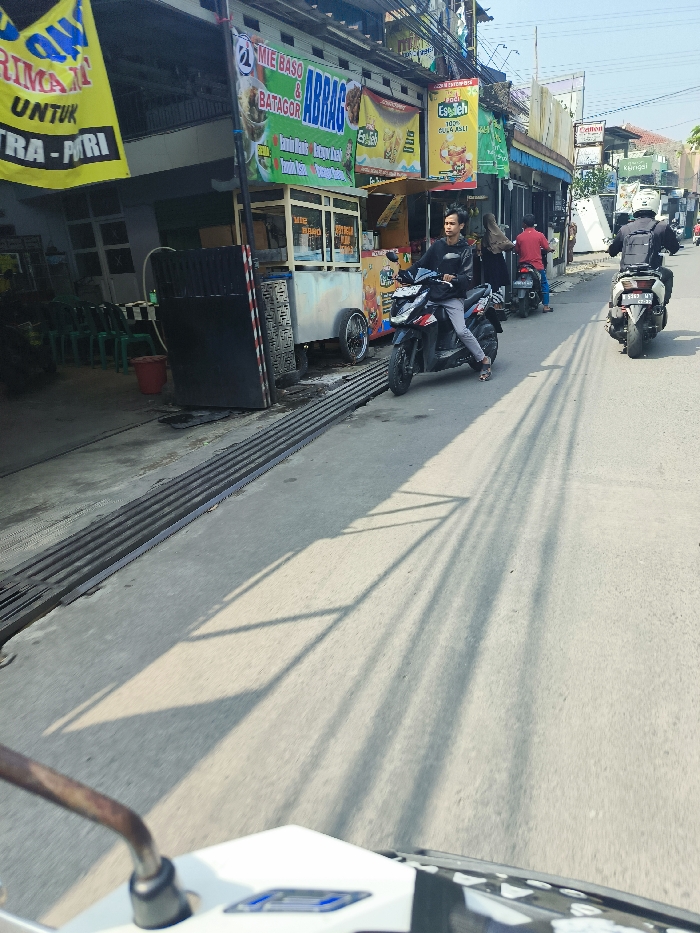 The image shows a street in Cileunyi, Indonesia (latitude: -6.94, longitude: 107.726). A man is driving a motorbike on the street, which is paved with asphalt. The man is wearing a black shirt and black pants. The motorbike is black and white. The street is lined with buildings on either side. There is a food stall on the left side of the street with a sign that says "Mie Baso & Batagor ABrag". The stall is selling snacks and drinks. There are shadows from trees on the ground. The person taking the photo is on a motorbike traveling behind the man on the motorbike. There are parked motorbikes and a person standing on the side of the street. The street has no pedestrians. The street is empty except for the motorbikes. The sky is blue and the sun is shining.