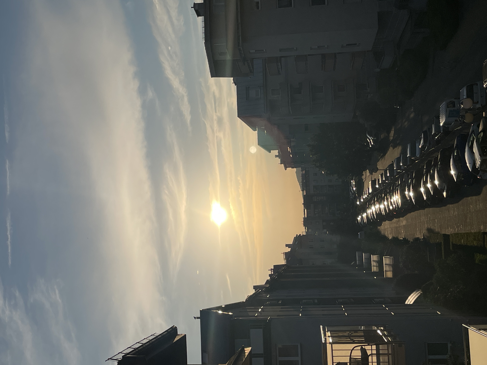 The image captures an urban scene at what appears to be dusk or dawn, given the low angle of the sun and the warm, golden hue of the sky. The sun is prominently visible, radiating light and casting long shadows. The sky is a mix of blue and yellow, with wispy clouds that are illuminated by the sunlight.
In the foreground, we see the rooftops and upper floors of several multi-story buildings. These buildings have a modern, somewhat stark architectural style, with visible windows and balconies. They are densely packed, suggesting an urban environment.
To the right of the image, a street or parking area is lined with numerous parked cars. The cars are reflecting the light from the sky, giving them a bright, metallic sheen. The vehicles are arranged in a row, indicating a parking lot or a street with parallel parking. Beyond the cars, more buildings and some trees are visible, contributing to the urban landscape.
The overall mood of the image is peaceful and serene, evoking the quiet transition between day and night. The low light and the long shadows create a sense of depth and atmosphere. There is no visible text in the image. The activity taking place is implied to be the end or beginning of a day in a city, with residents likely in their homes or the street scene of parked vehicles.
