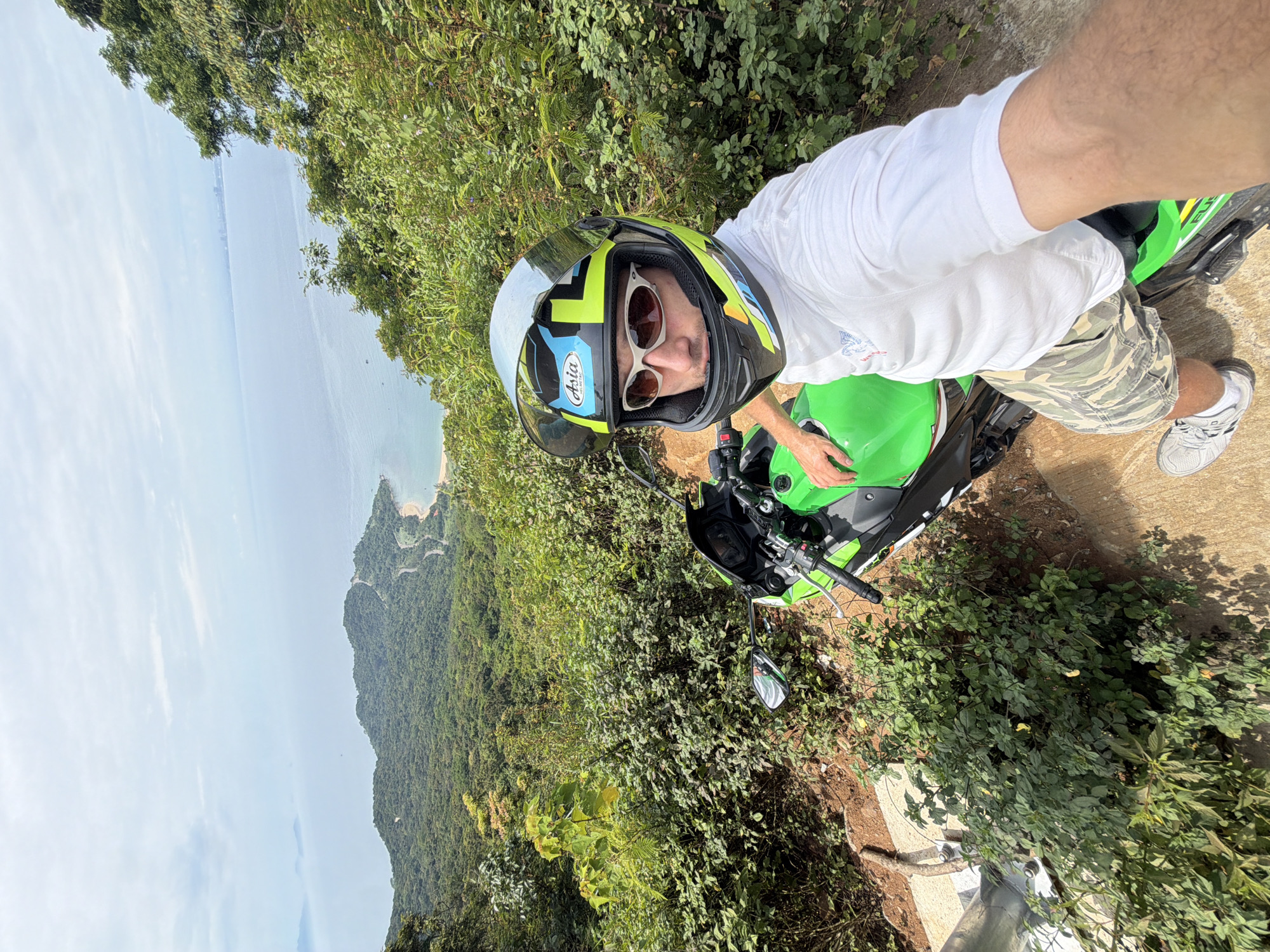 A man on a green Kawasaki Ninja motorcycle takes a selfie on a roadside overlook, likely on the Sơn Trà Peninsula, Đà Nẵng, Vietnam. He wears a motorcycle helmet, sunglasses, a white t-shirt, and camouflage shorts. The background shows a coastline with a bay, forest-covered hills, and the sea. The sky is overcast. The photograph is taken at an angle, with the motorcycle and the man occupying the foreground and the coastal view in the background. The roadside is lined with shrubs and trees.