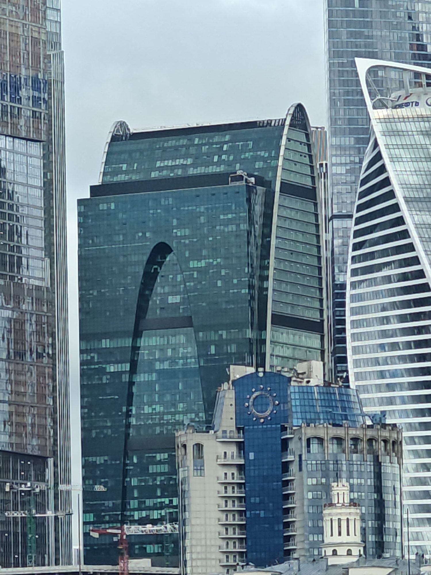 This is a photograph of the Moscow International Business Center, also known as Moscow-City, in Moscow, Russia. The image is a close-up view of several modern skyscrapers, predominantly featuring glass facades.
In the foreground, a large building with a distinctive arched opening in its glass facade is prominent. This building appears to be dark teal or blue in color. To the left of this structure is another tall skyscraper with a glass facade, reflecting the sky and surrounding buildings. To the right, a white, triangular-shaped skyscraper dominates the background, with horizontal white stripes along its facade. Partially visible behind these structures are other skyscrapers with similar modern architectural styles.
Below the main skyscrapers, a section of a building with a blue glass facade and a circular decorative element resembling a clock face is visible. This is flanked by lower buildings with multiple windows and balconies, suggesting a mixed-use development. The sky is overcast, contributing to the cool, diffused lighting of the scene. No people are visible in the image. The overall impression is one of a dense, contemporary urban landscape characterized by impressive architectural designs.