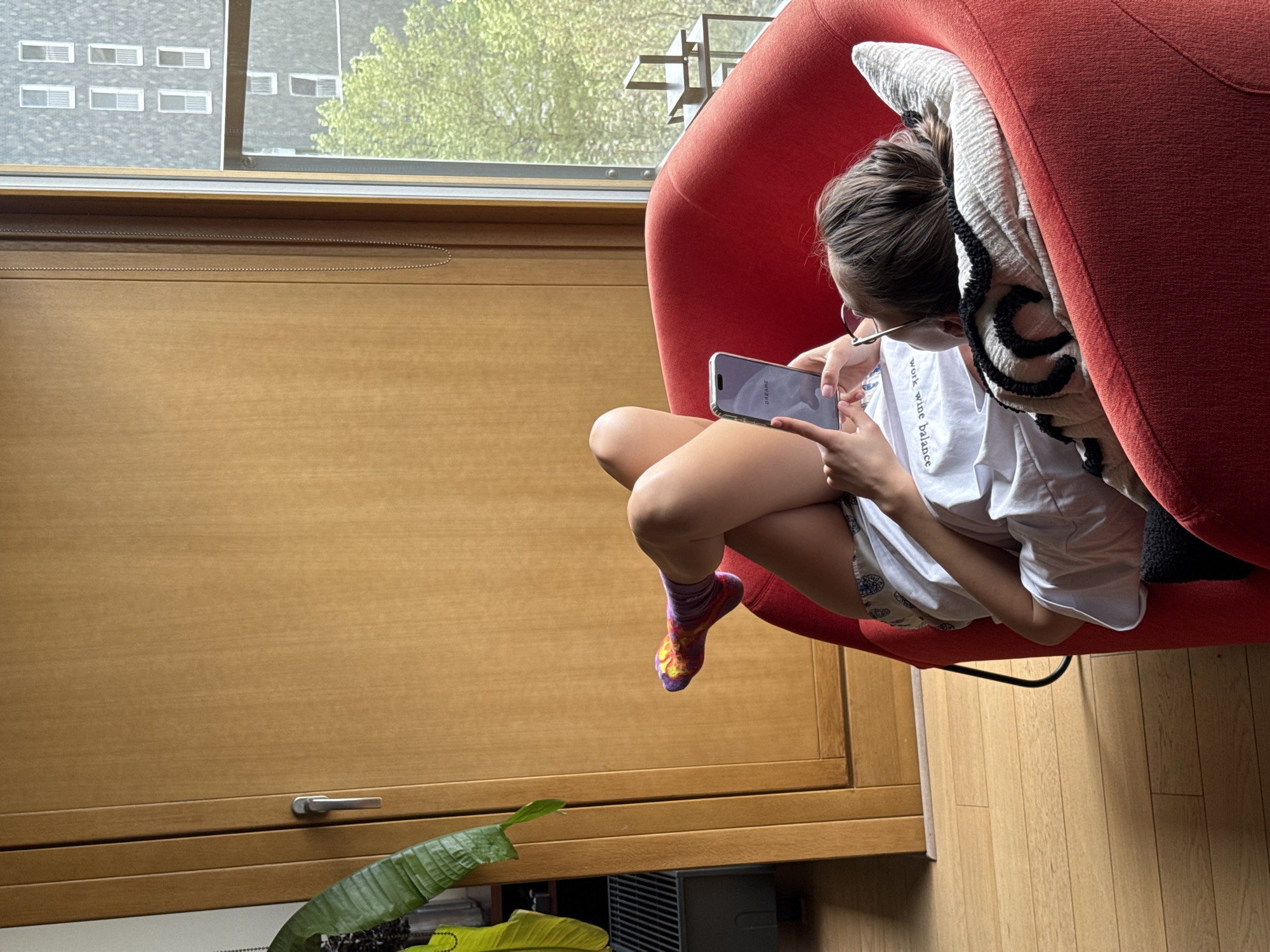 A young person wearing glasses and colorful socks is sitting in a red, rounded chair near a window, looking at a smartphone. The person is wearing a white t-shirt and patterned shorts. A plant is visible near a wooden cabinet. The location appears to be inside a residence.